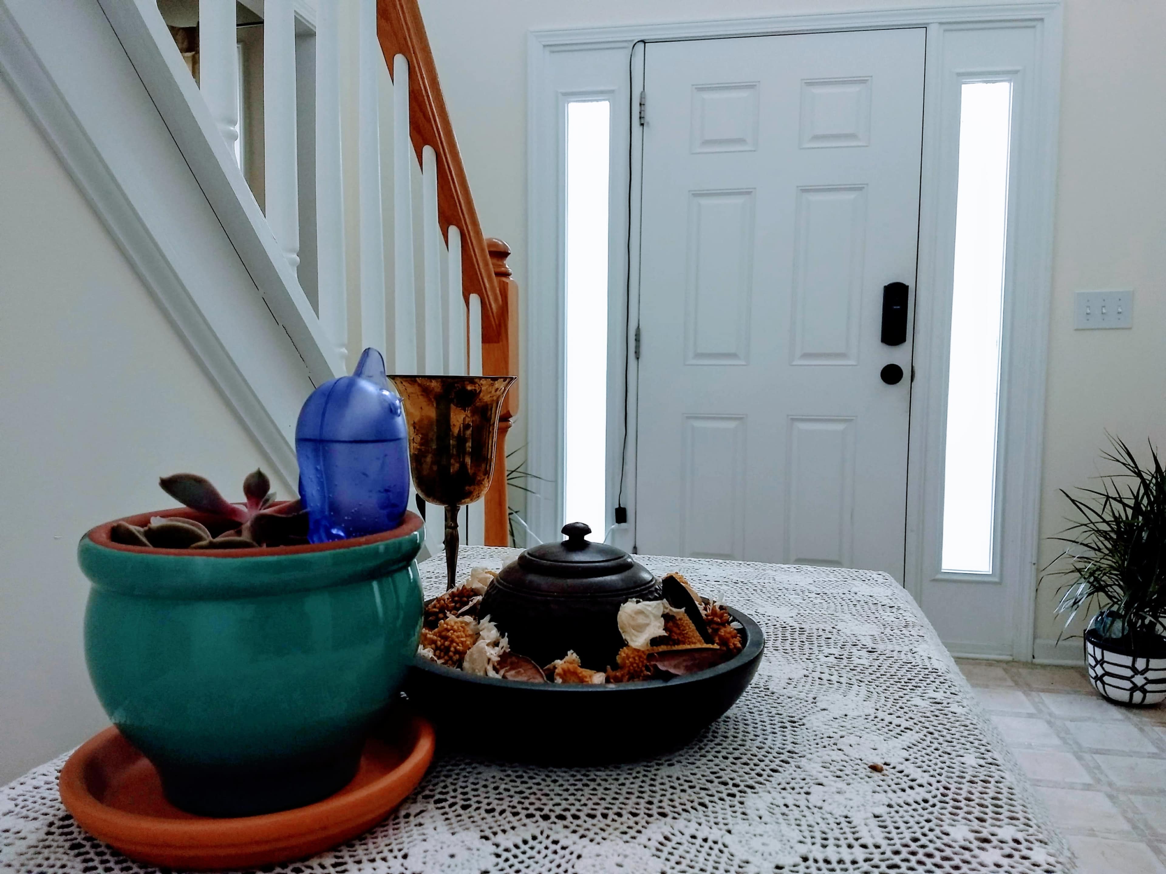 A table is set in an entryway, featuring a turquoise pot with plants, a blue water bottle, a decorative bowl filled with snacks, and a decorative cup, with a staircase and front door visible in the background.