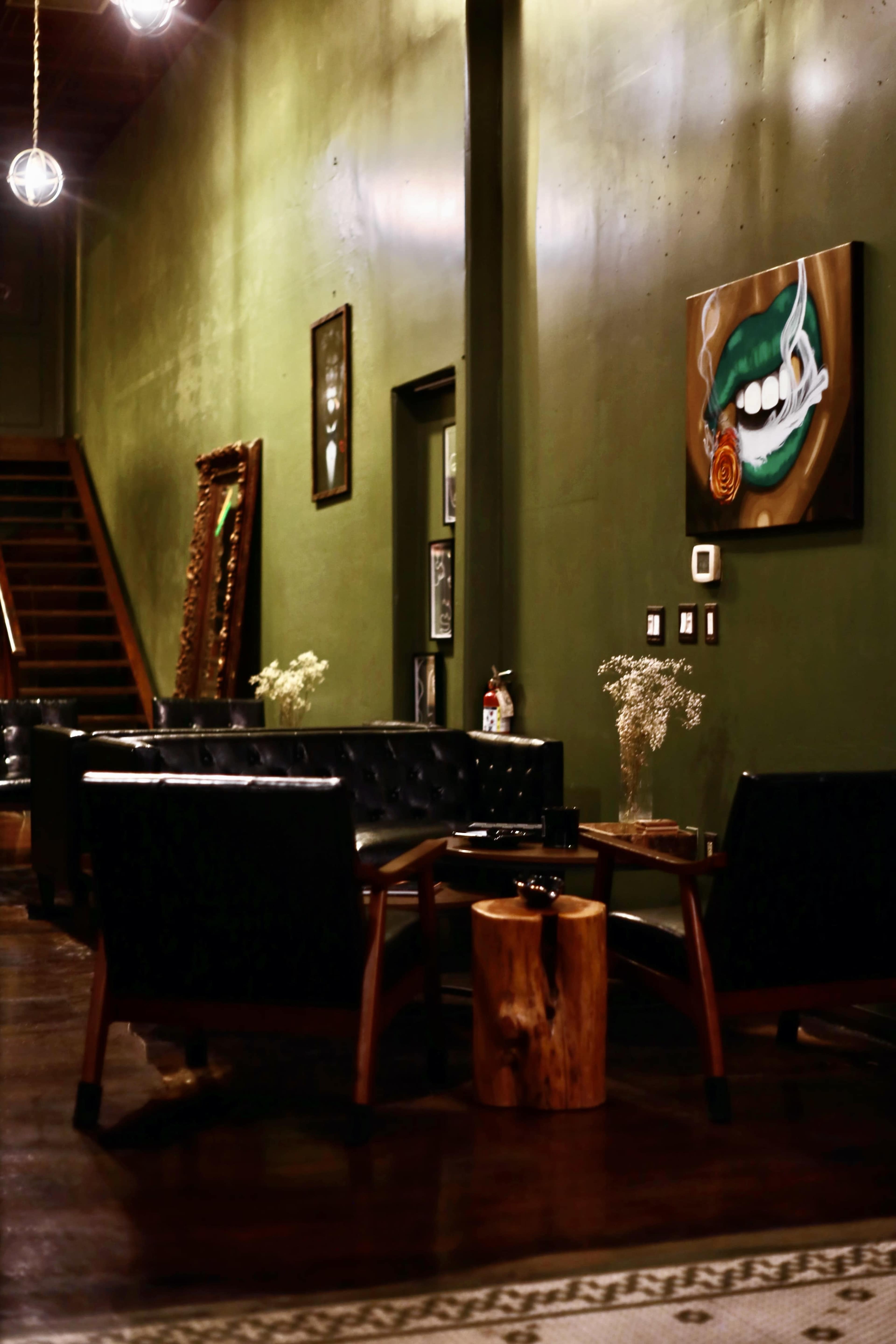 The image shows a green-walled lounge area with black leather chairs, a wooden coffee table, and a painting of a mouth with a smoking cigarette.