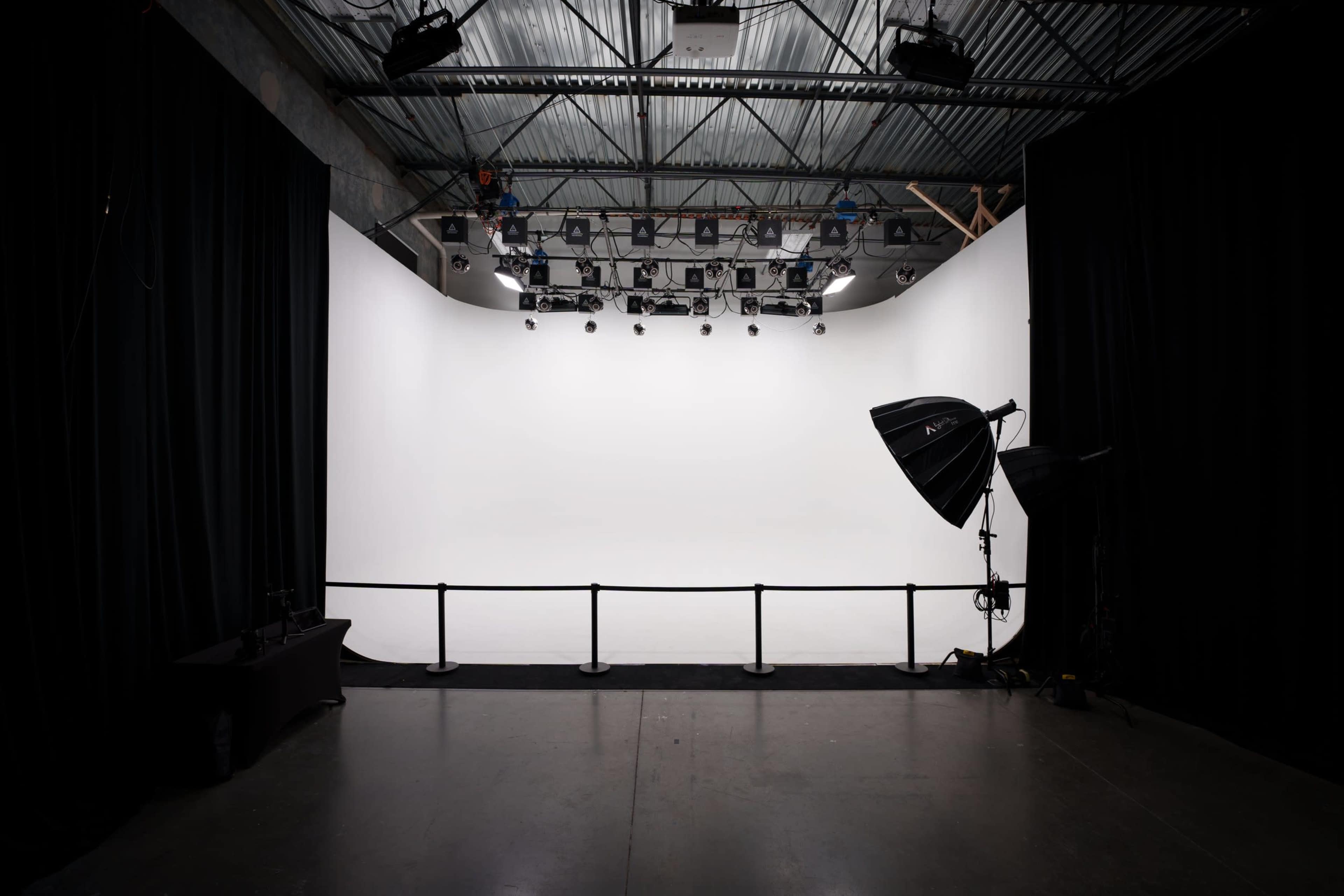 A photography studio features a large, empty white backdrop with lighting equipment and a railing in the foreground.