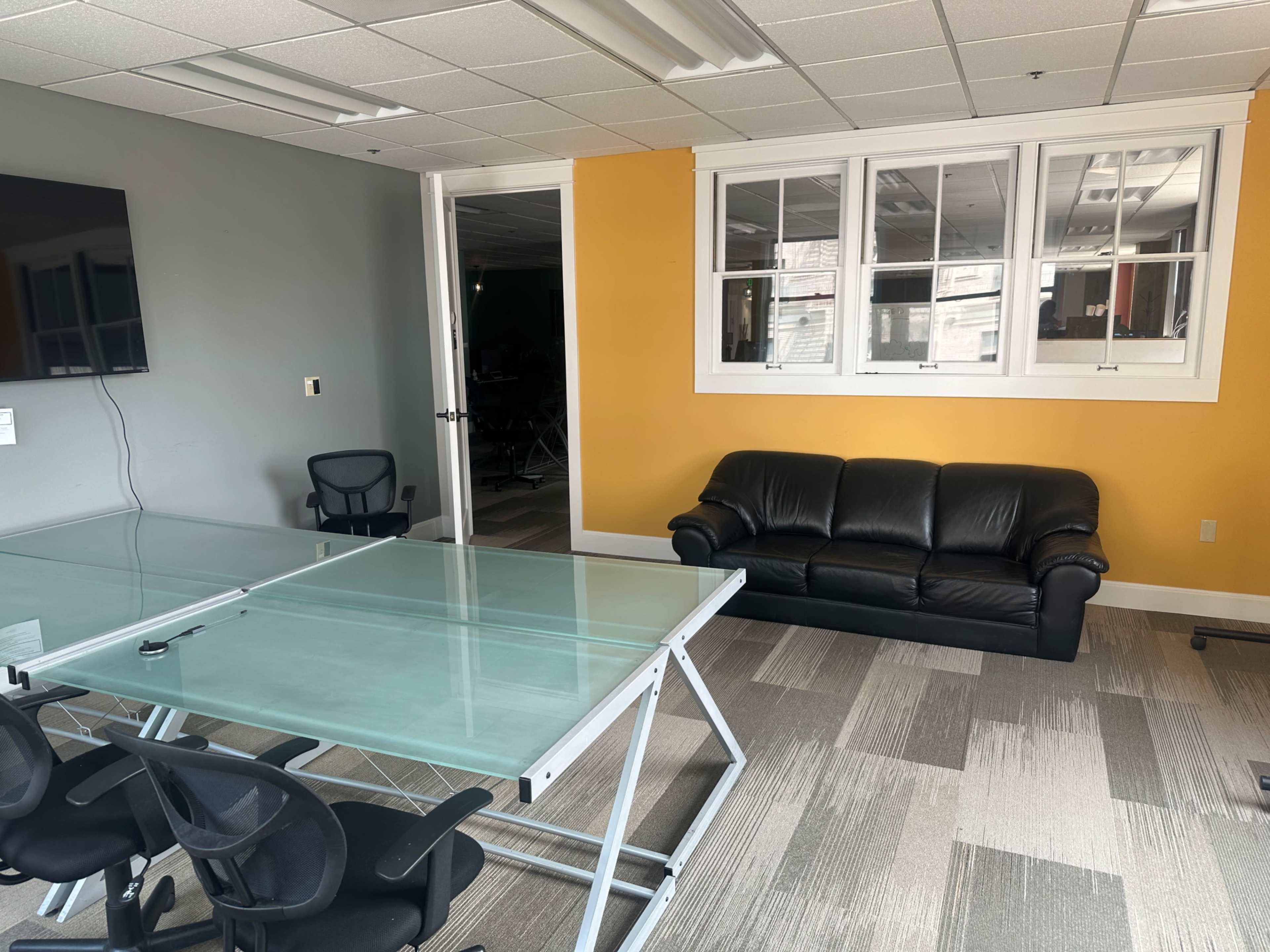 The image shows a conference room with a glass table, black ergonomic chairs, and a leather sofa against an orange wall, along with a television mounted on the wall.