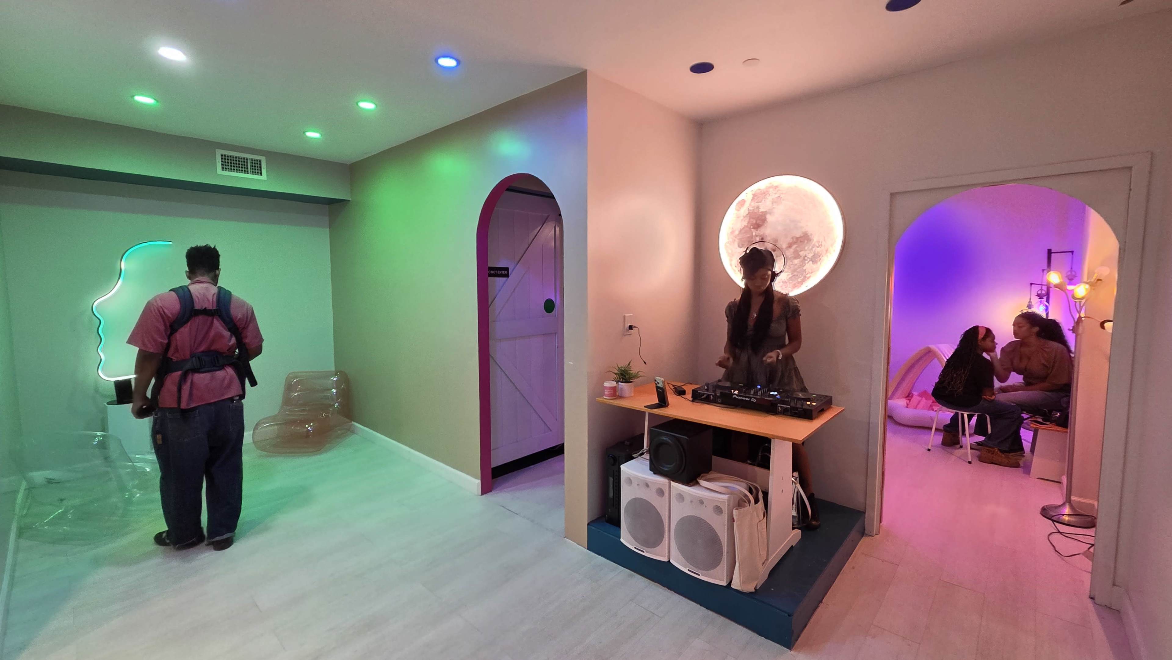 The image shows a brightly colored room divided into two areas: one with a person standing near a translucent installation and the other featuring a DJ playing music in front of a circular light.