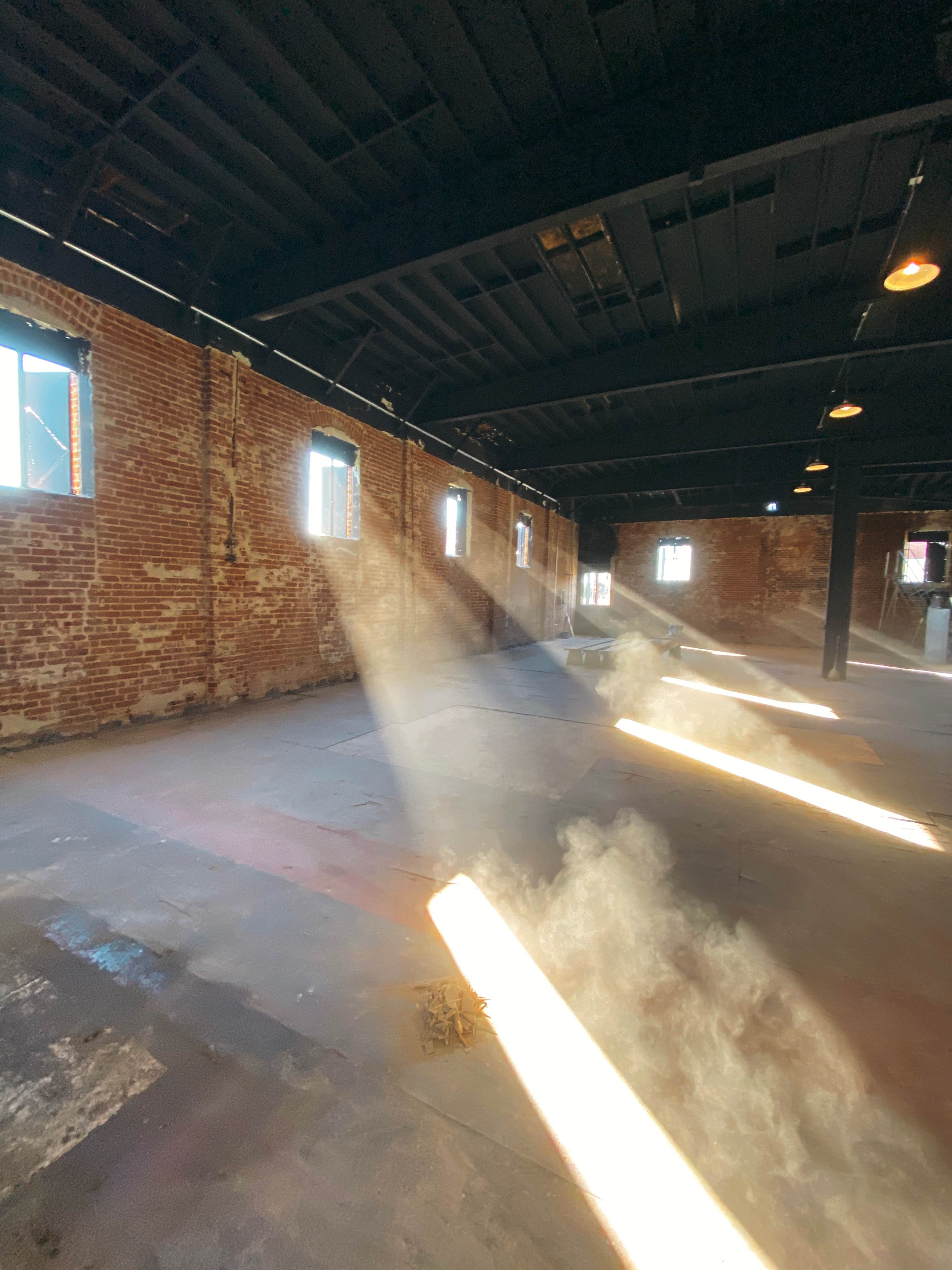 Sunlight streams through windows, creating beams that illuminate the dust in an empty, brick-walled warehouse.