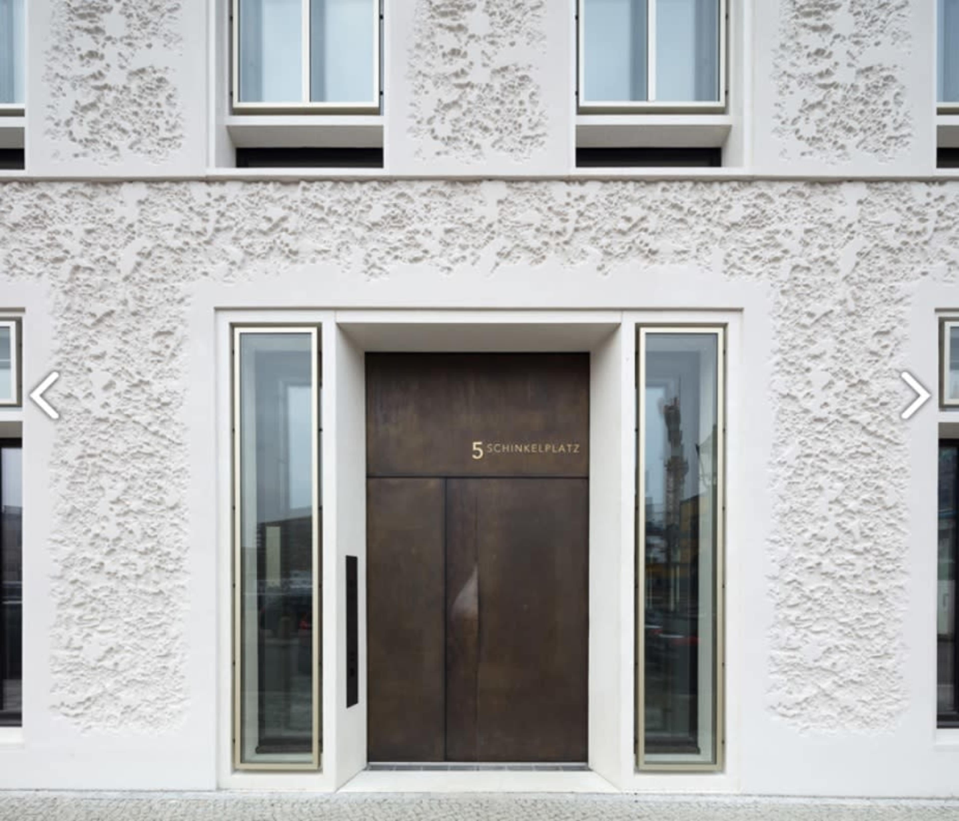A modern building facade featuring a large, dark wooden door with the number "5" and the word "SCHINKENPLATZ" above it, framed by large glass panels.