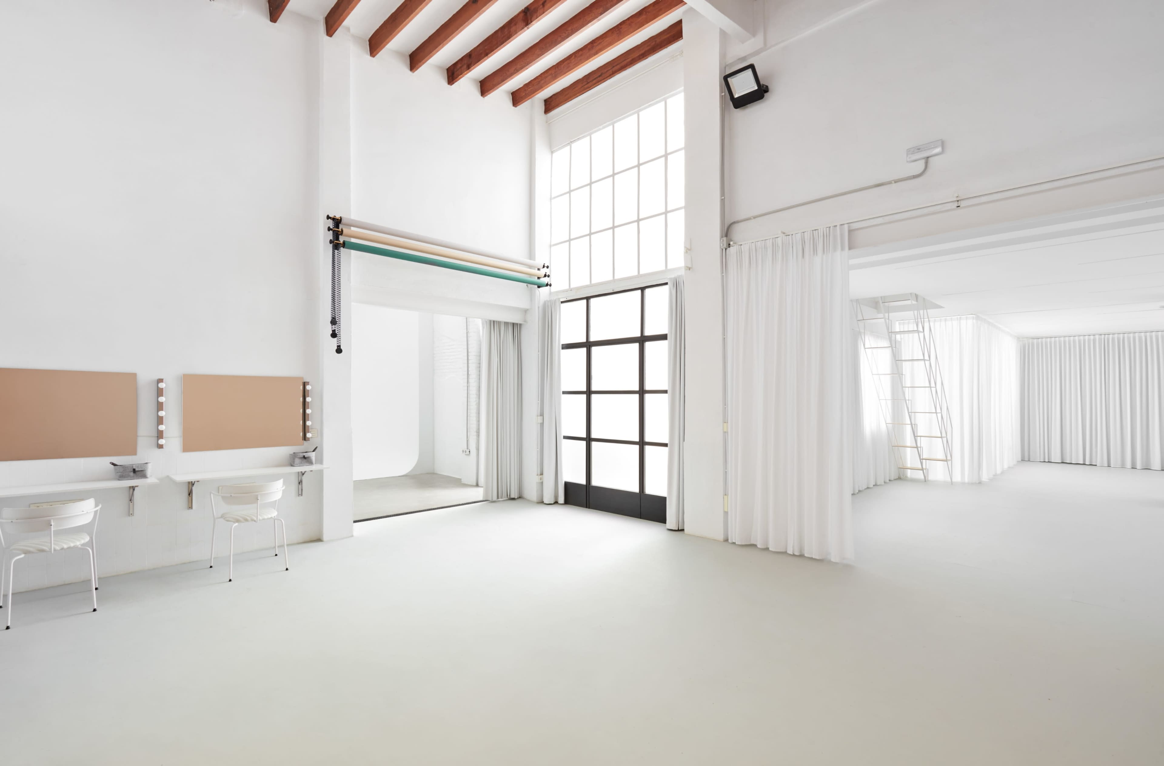 The image depicts a spacious, well-lit studio with white walls, a large window, and two vanity stations with mirrors.