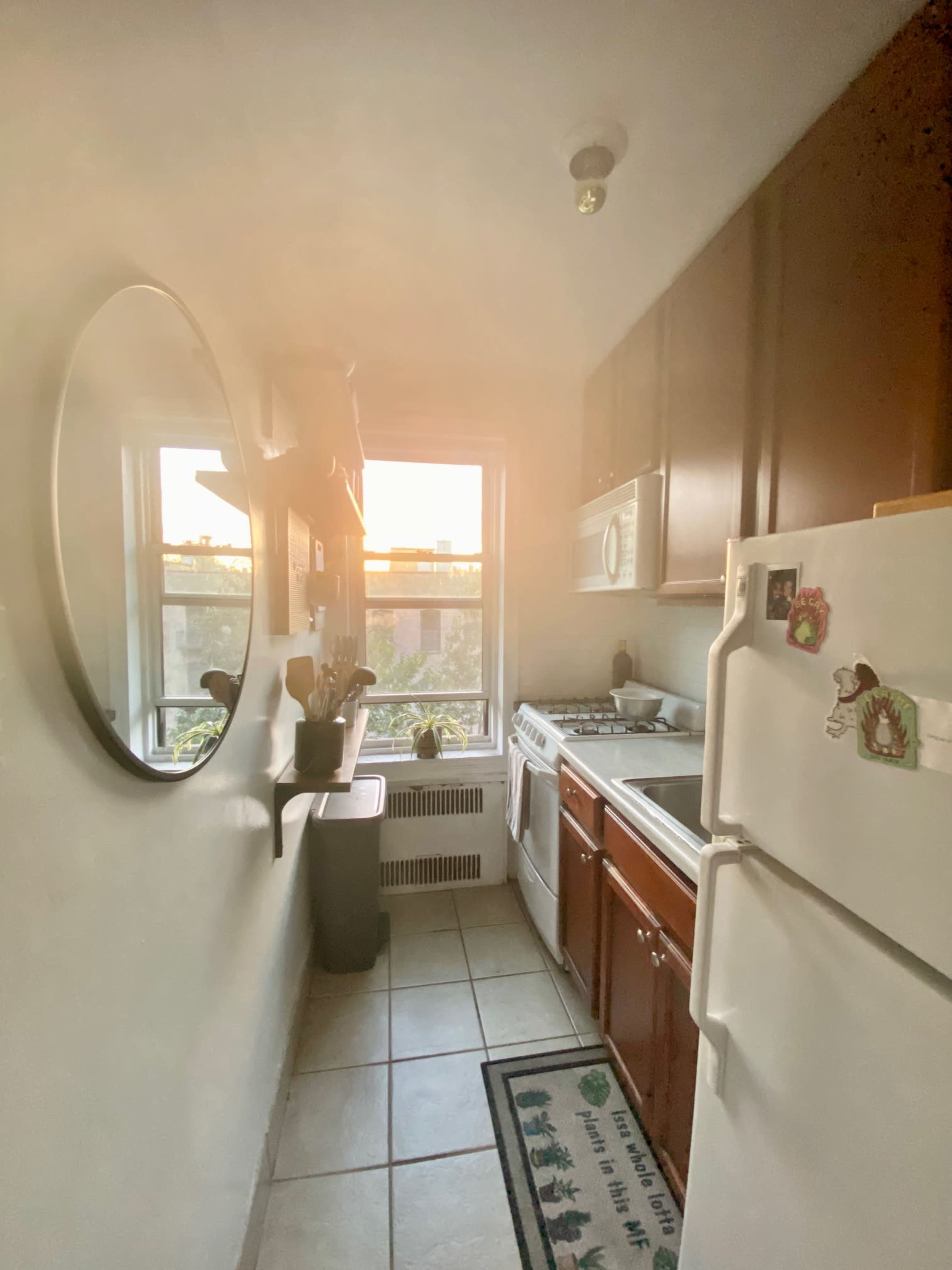 A small kitchen features a window with sunset light, a stove, a refrigerator, and a mirror on the wall.
