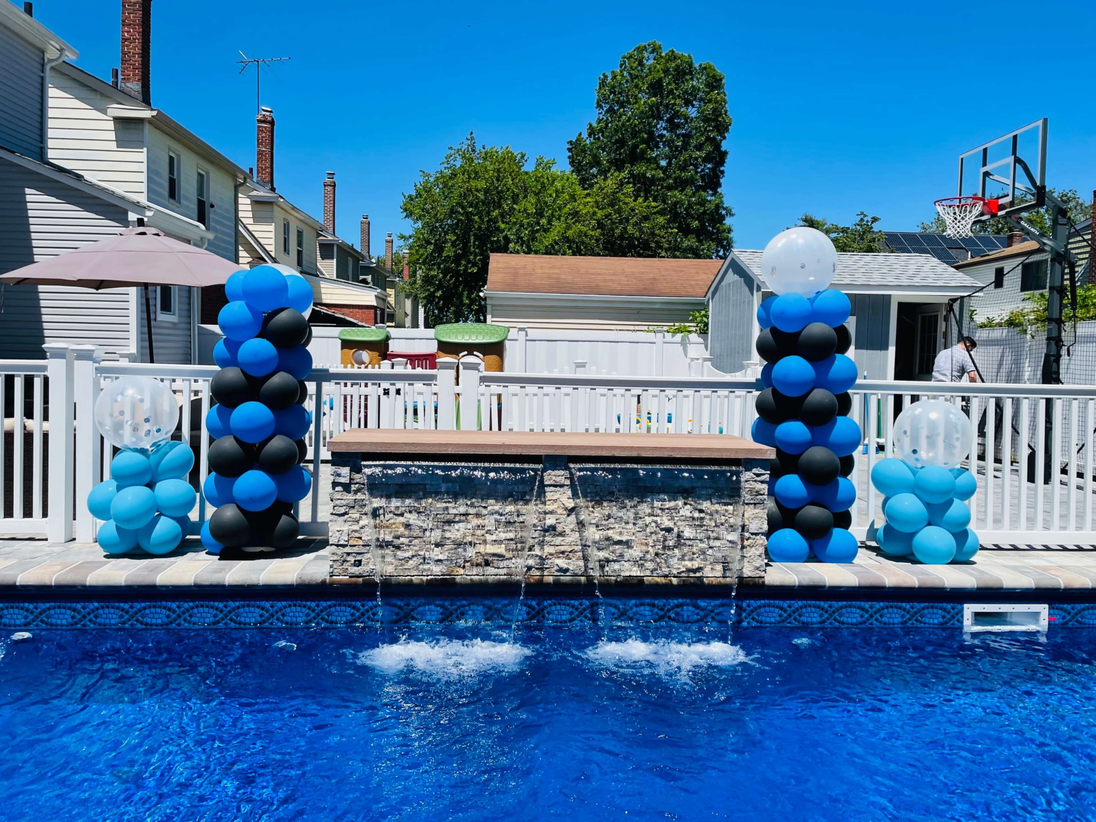 The image shows a swimming pool area decorated with blue and black balloon columns on either side of a stone water feature, under a clear blue sky.
