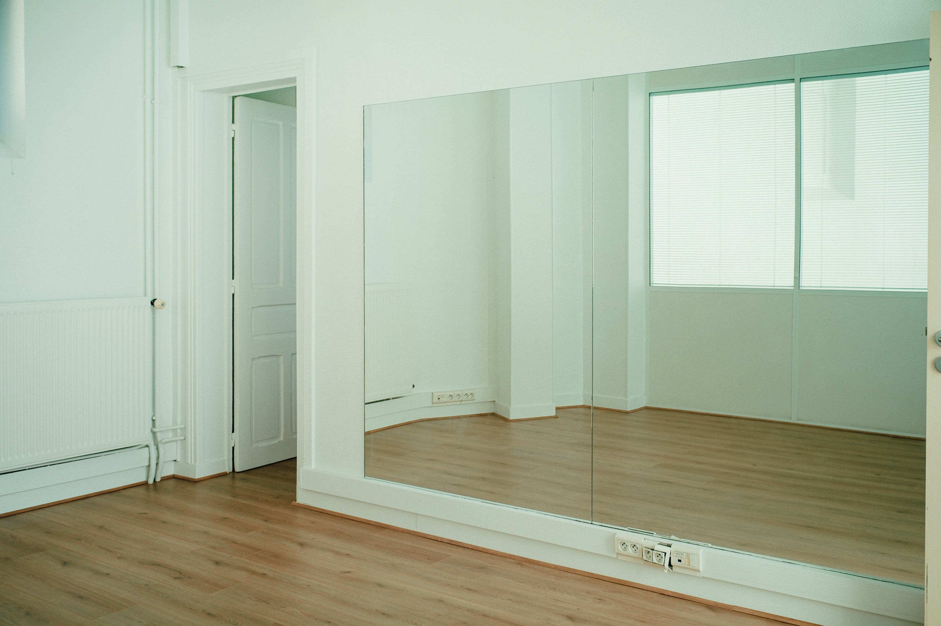 A vacant room with a mirror wall and a wooden floor, featuring a door and a window with vertical blinds.