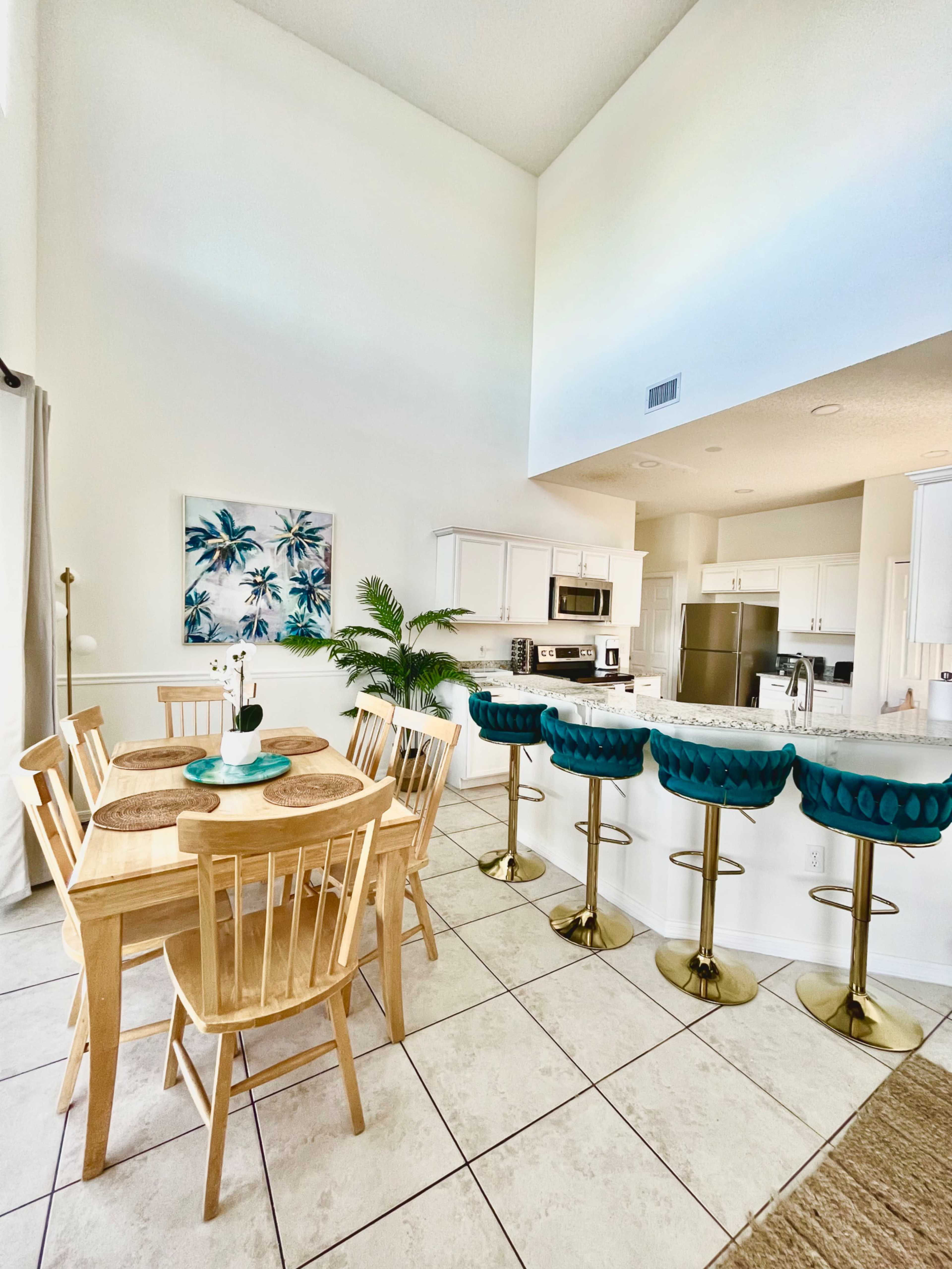 A modern kitchen and dining area feature a wooden table with four chairs, turquoise bar stools at a counter, and a decorative plant.