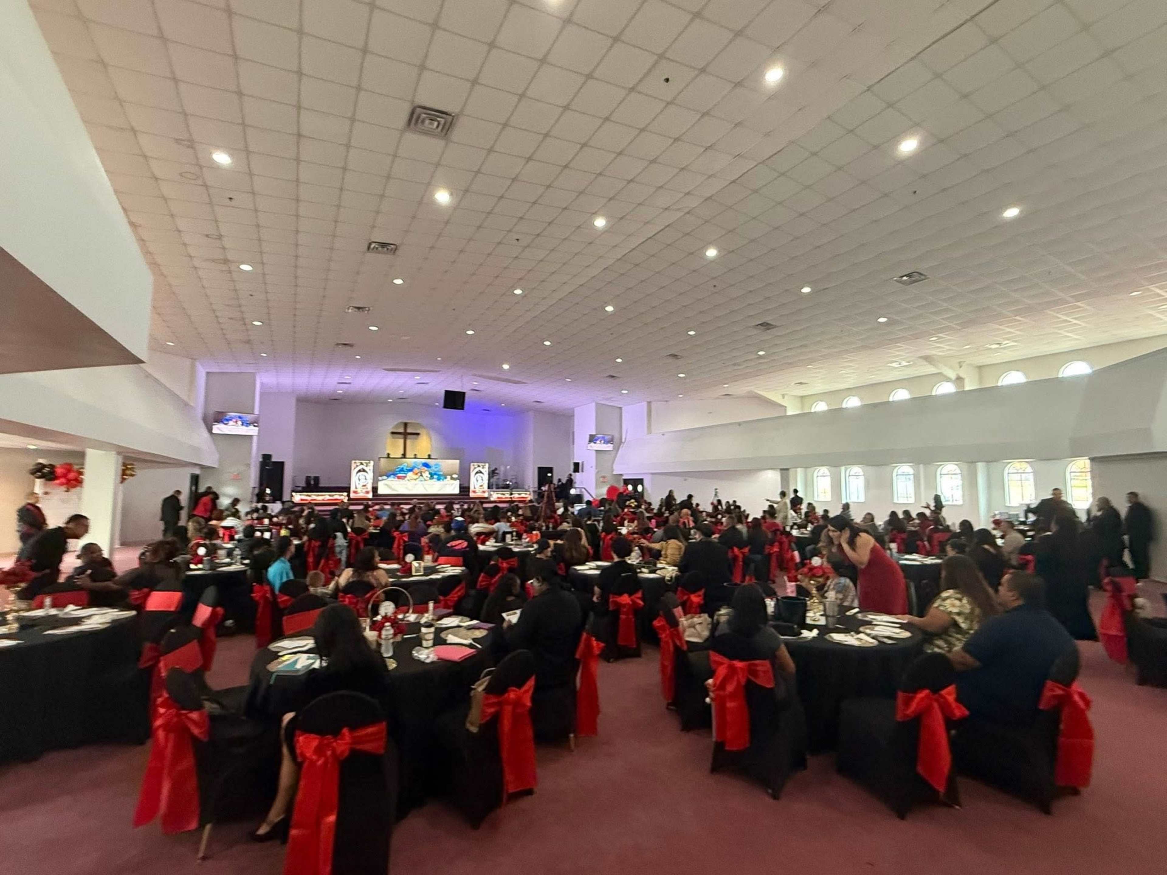 A large banquet hall is filled with guests seated at tables adorned with black cloths and red bows, with a stage set up in the background.