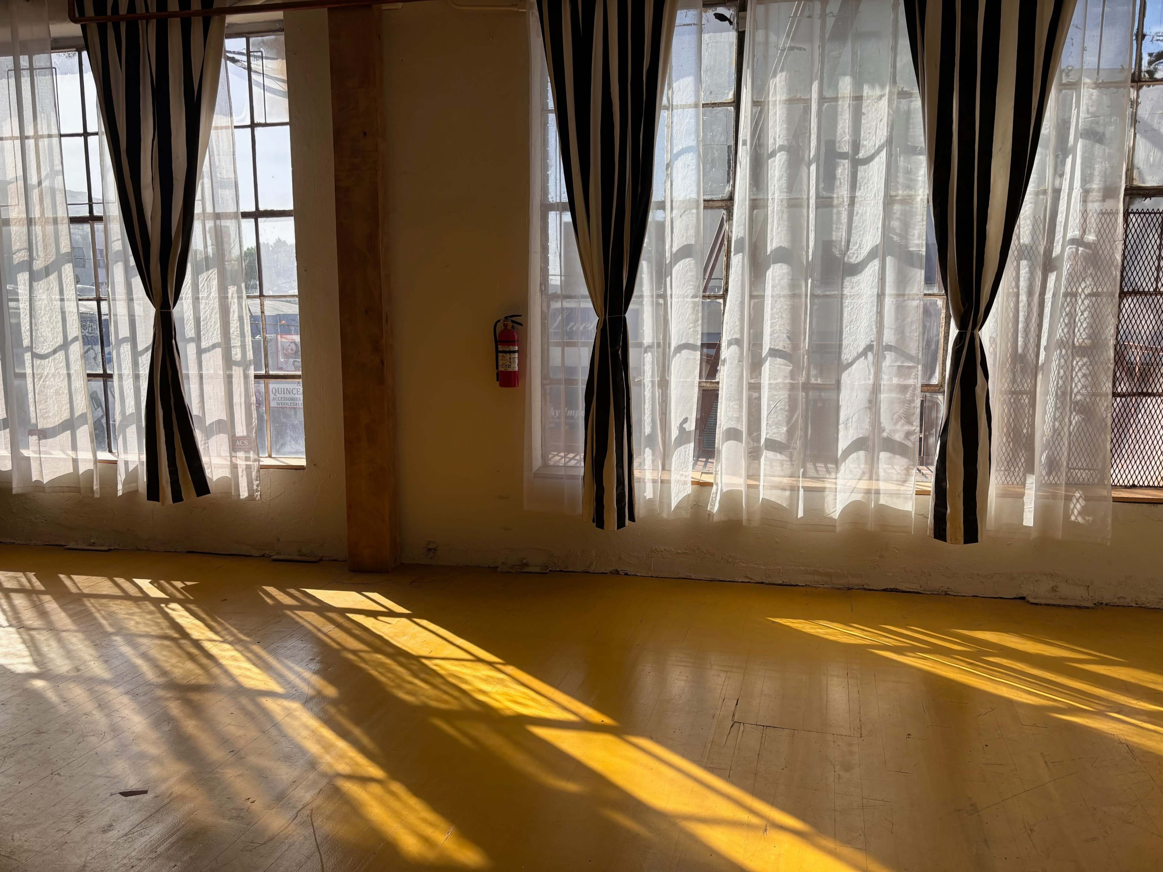 Sunlight filters through sheer curtains, casting striped shadows on a yellow wooden floor in a spacious room.