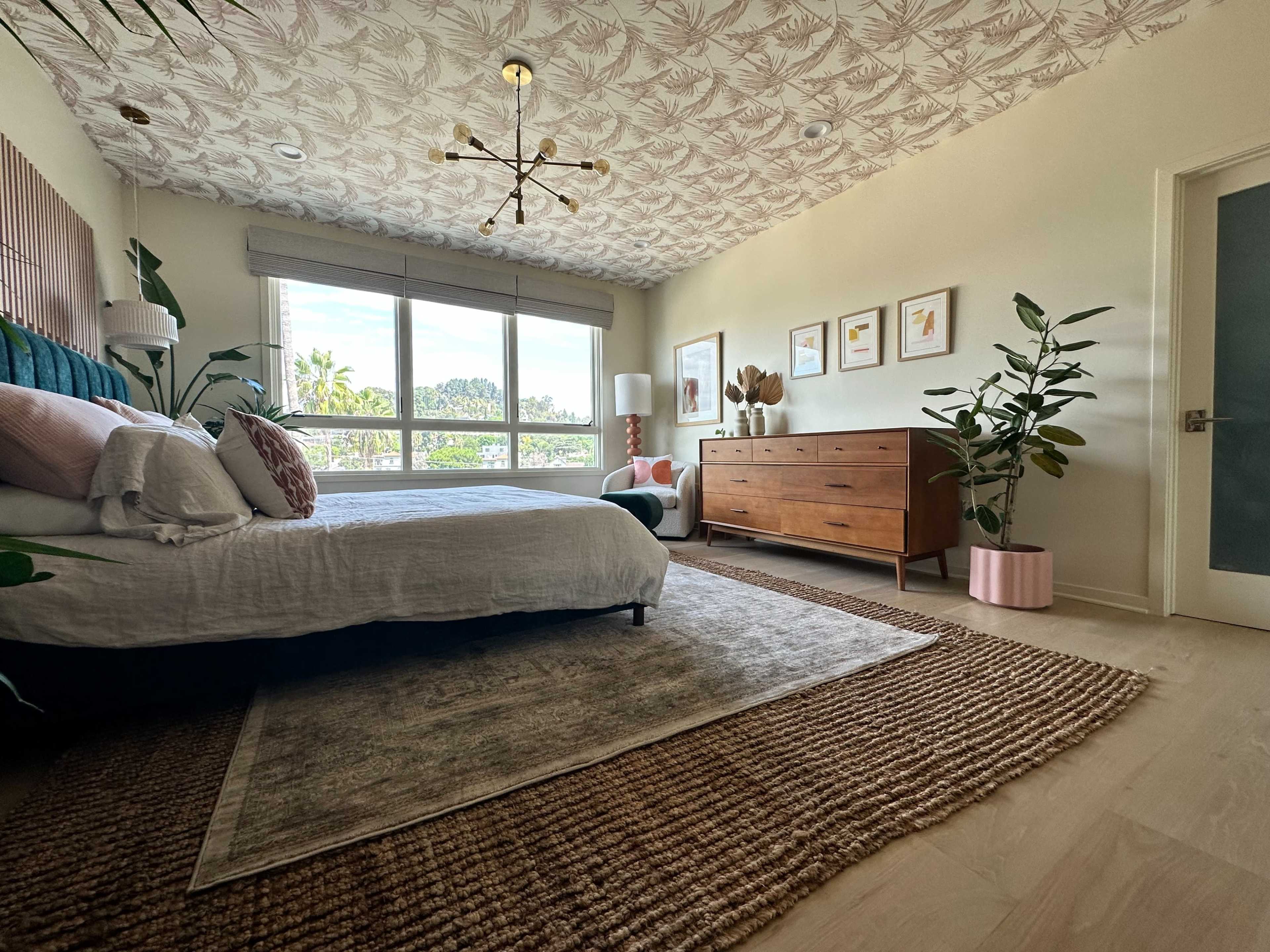 A modern bedroom with a large window, a bed with decorative pillows, a wooden dresser, a patterned ceiling, and several plants.