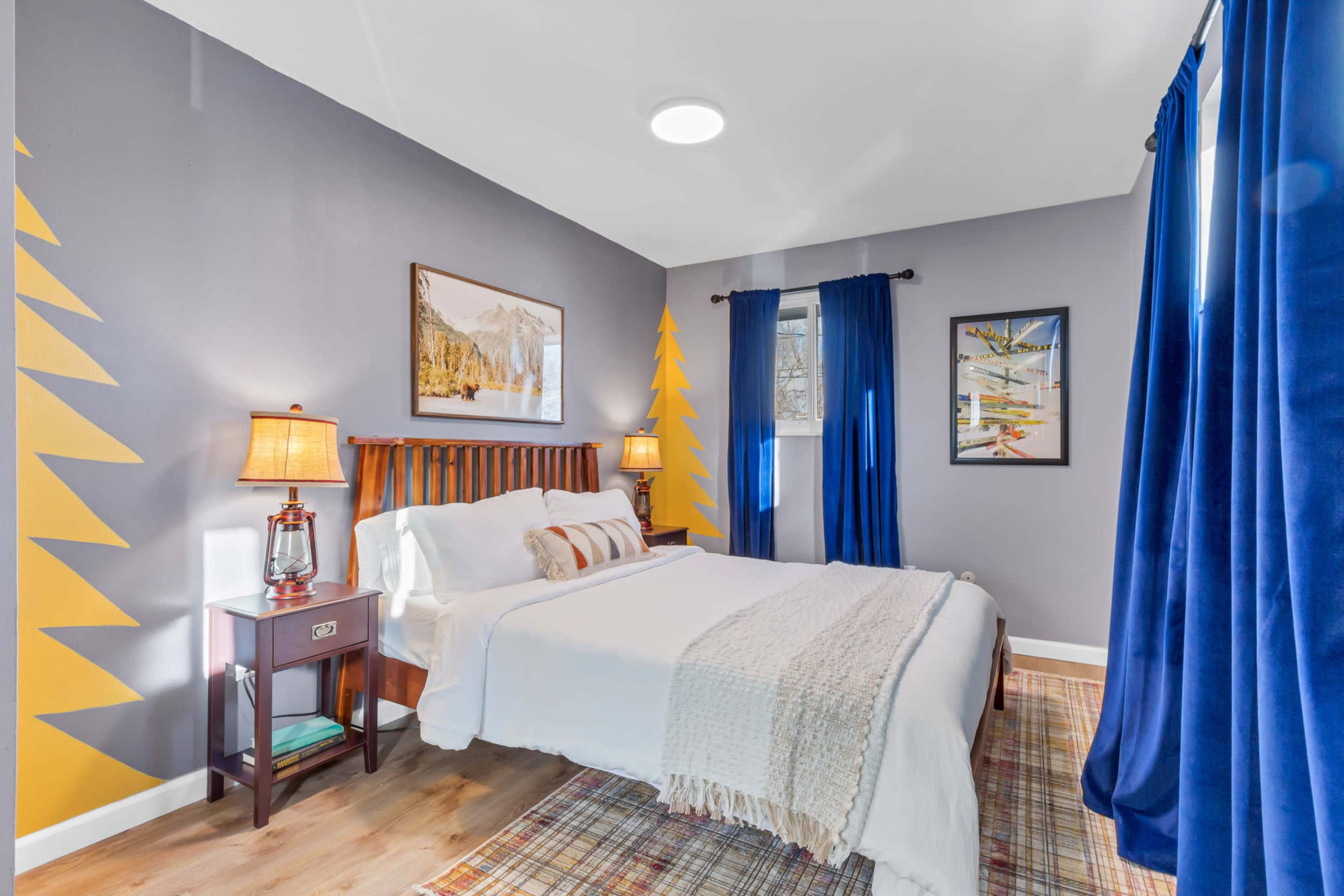A cozy bedroom featuring a bed with white linens, a wooden headboard, yellow accent wall designs of trees, and dark blue curtains.