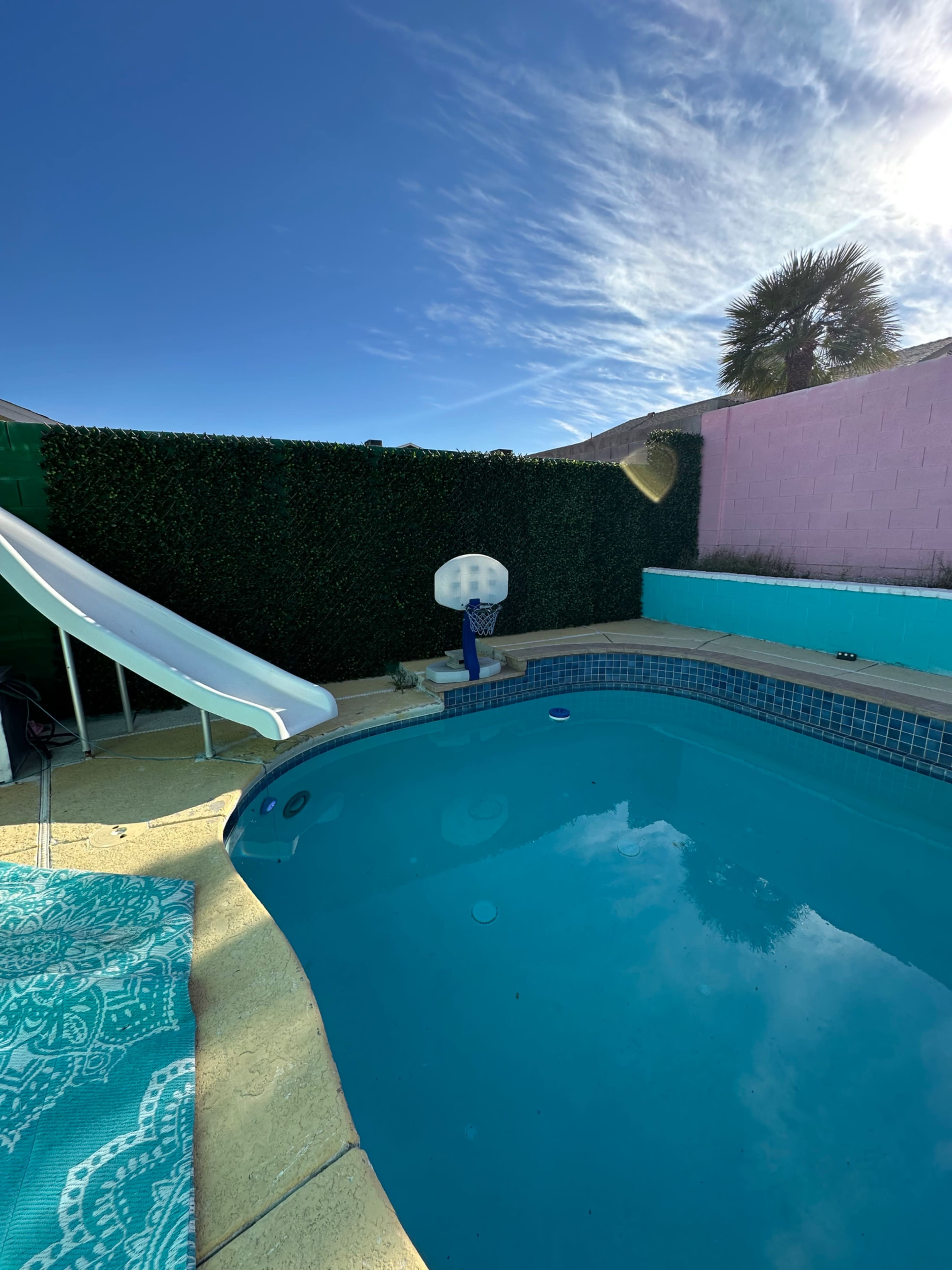 The image shows a swimming pool with a slide on one side, surrounded by a green hedge and a pink wall under a clear blue sky.