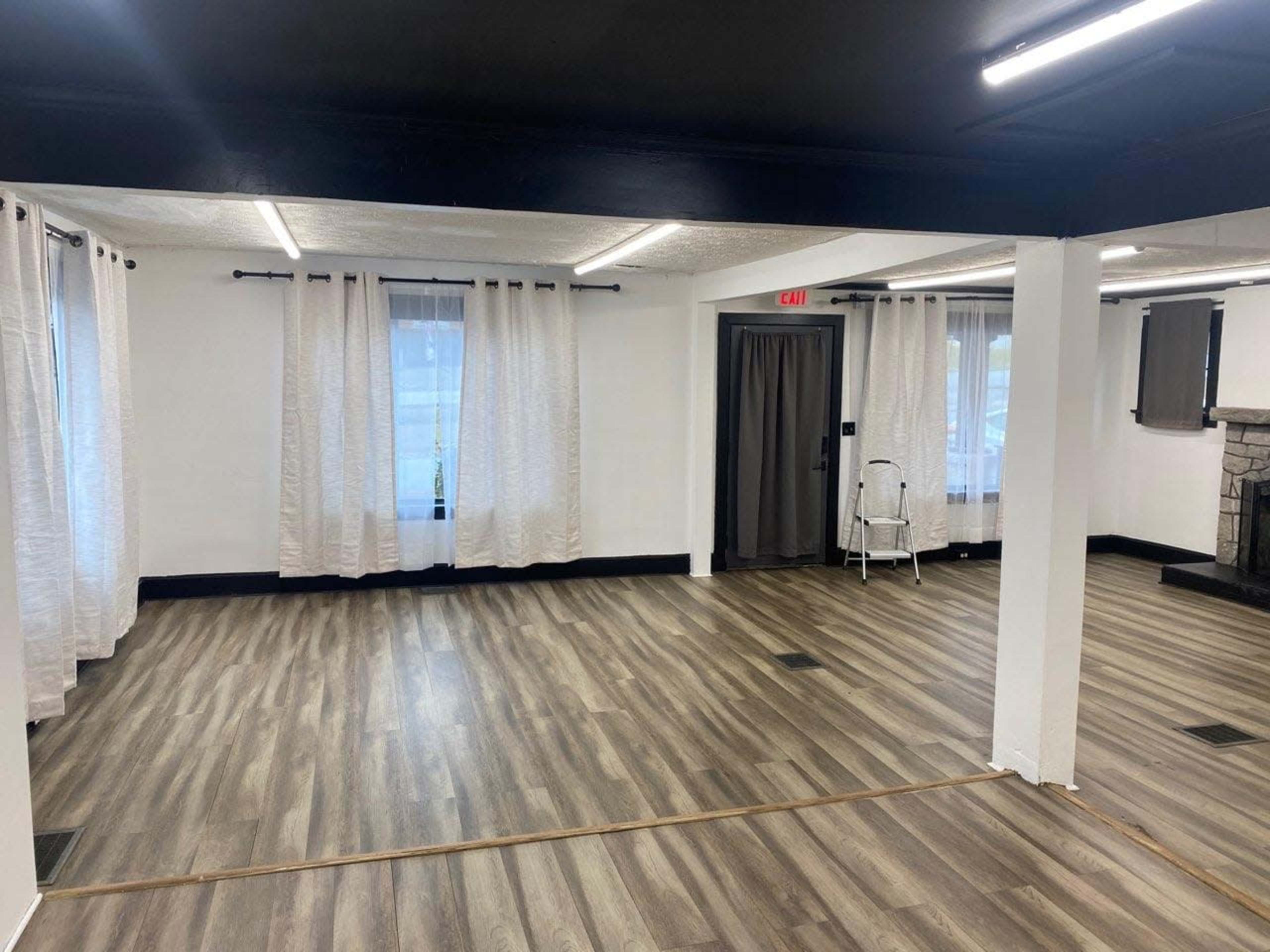 The image shows a spacious room with light-colored wooden flooring, white curtains on the windows, and minimal furniture, including a folding chair.