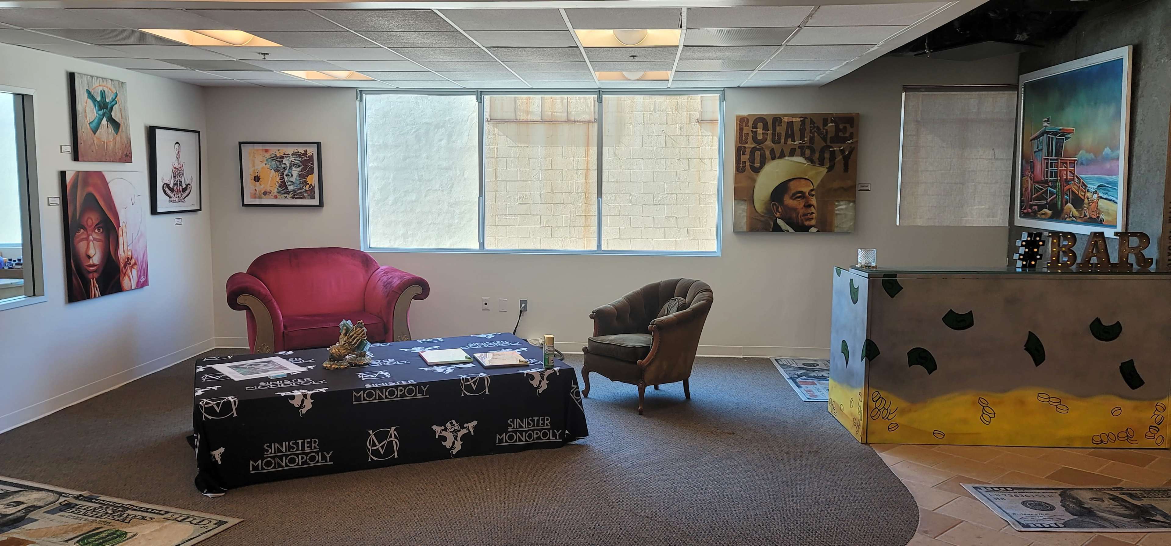 A modern lounge area with a pink sofa, a brown armchair, and a table covered in a black tablecloth featuring a Monopoly design, alongside wall art and a bar counter.
