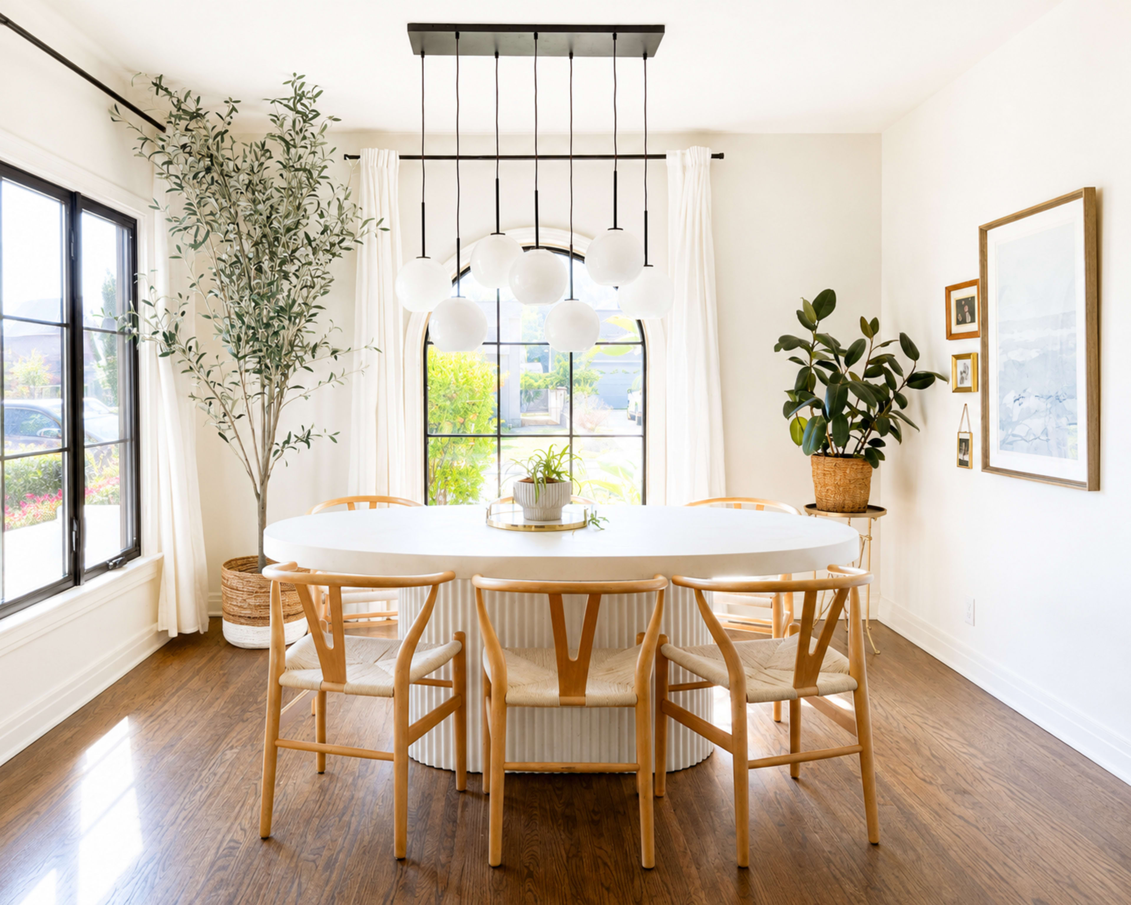 A modern dining room features a large oval table surrounded by wooden chairs, with a statement light fixture above and plants positioned near large windows.