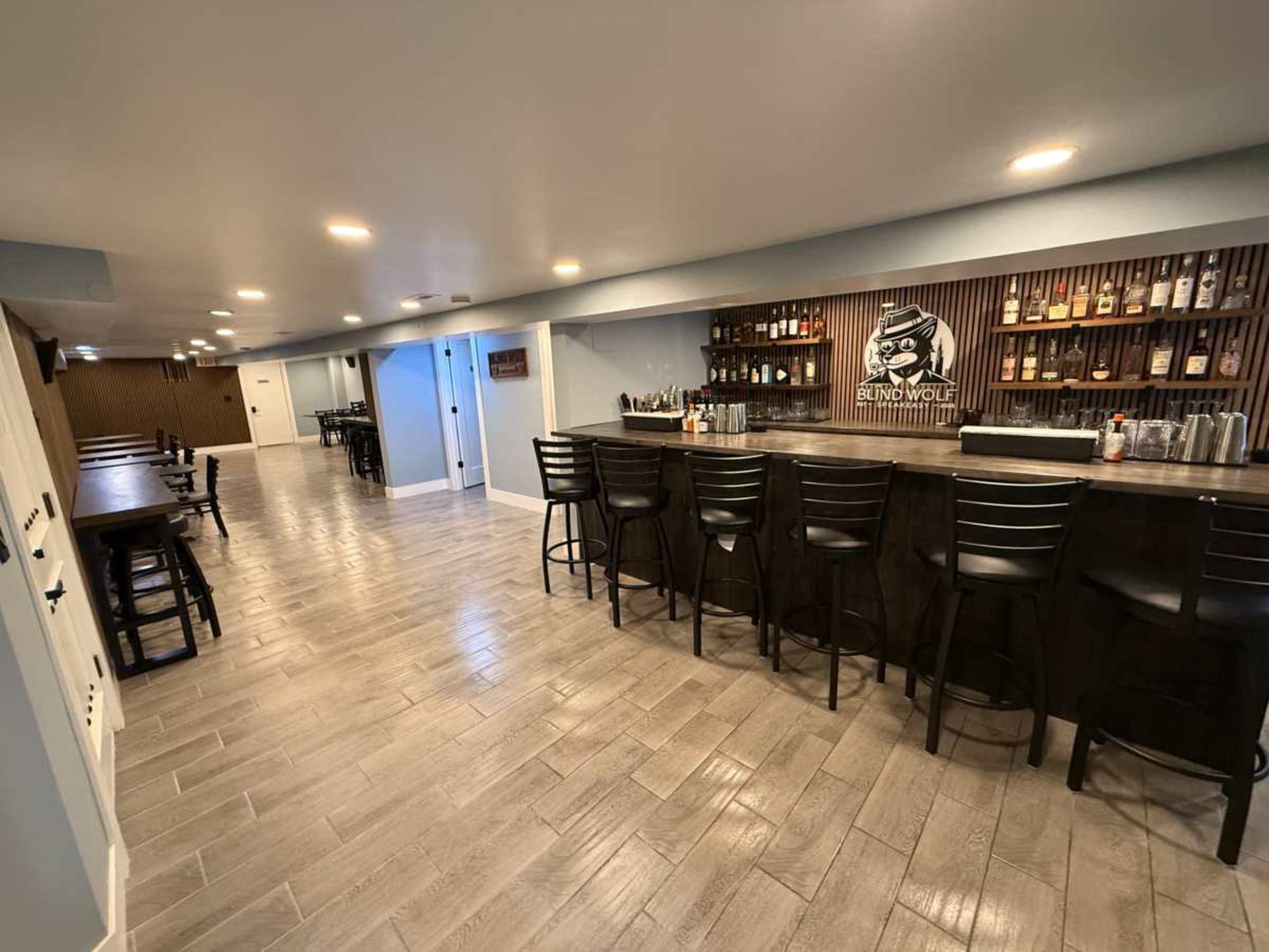 The image shows a spacious basement bar area with a long counter, high chairs, and a wooden wall displaying various bottles of liquor.