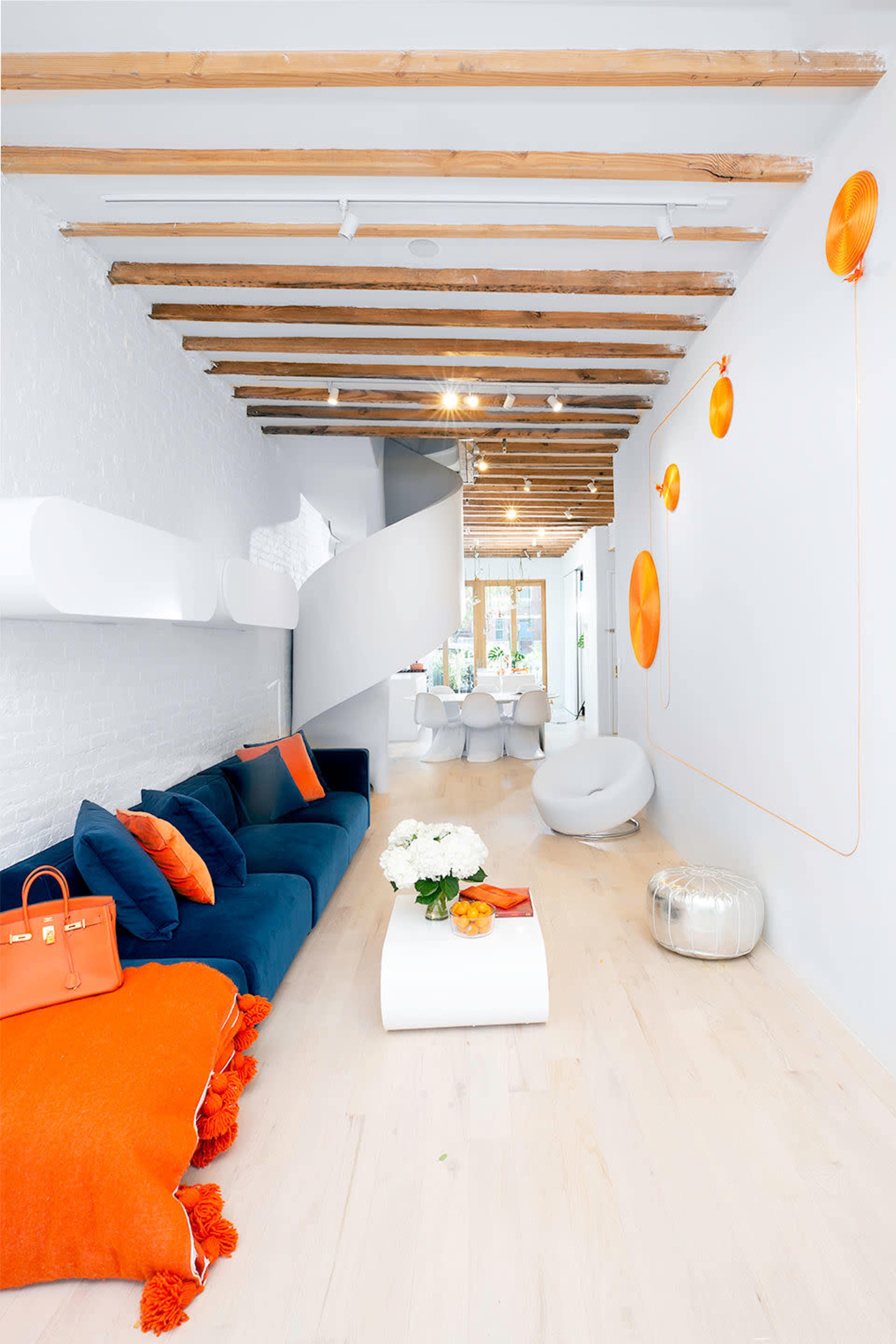 A modern living space features a blue sofa with orange accents, streamlined furniture, and exposed wooden beams overhead.