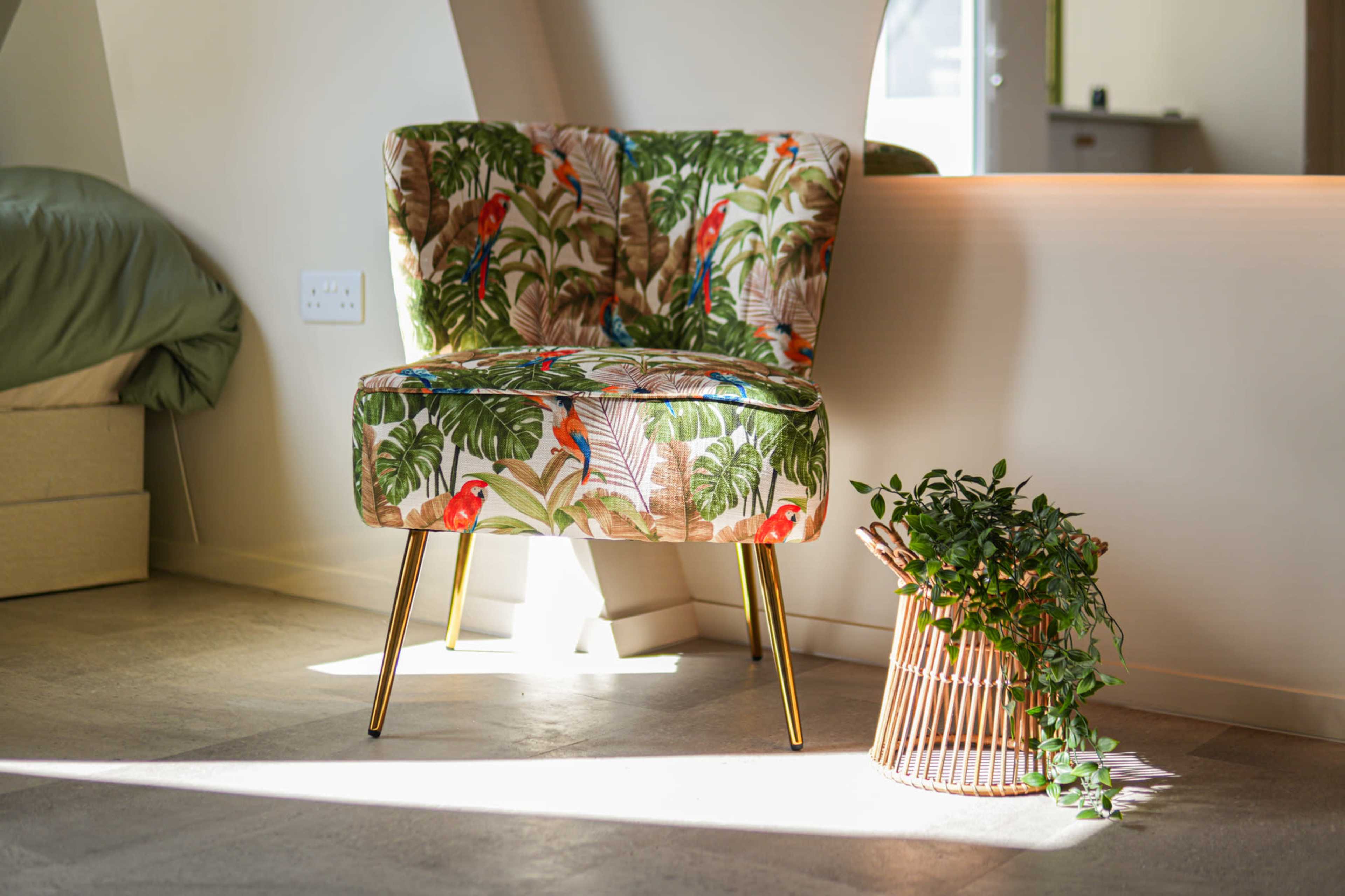 A floral-patterned chair with gold legs sits beside a woven basket containing green plants in a well-lit interior space.