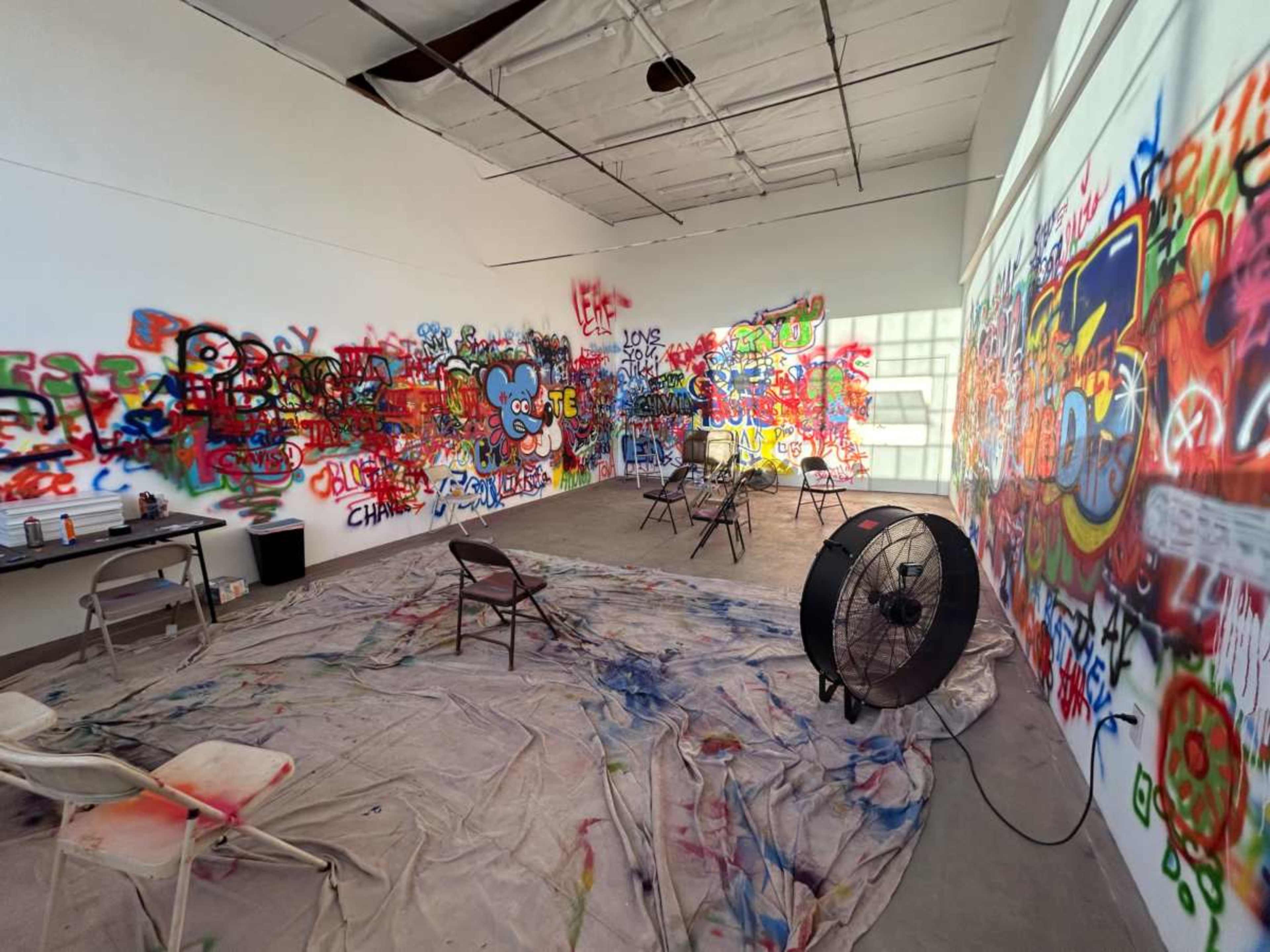 A room with various colorful graffiti on the walls, a few folding chairs, and a large fan on the floor amidst a tarp covered in paint splatters.