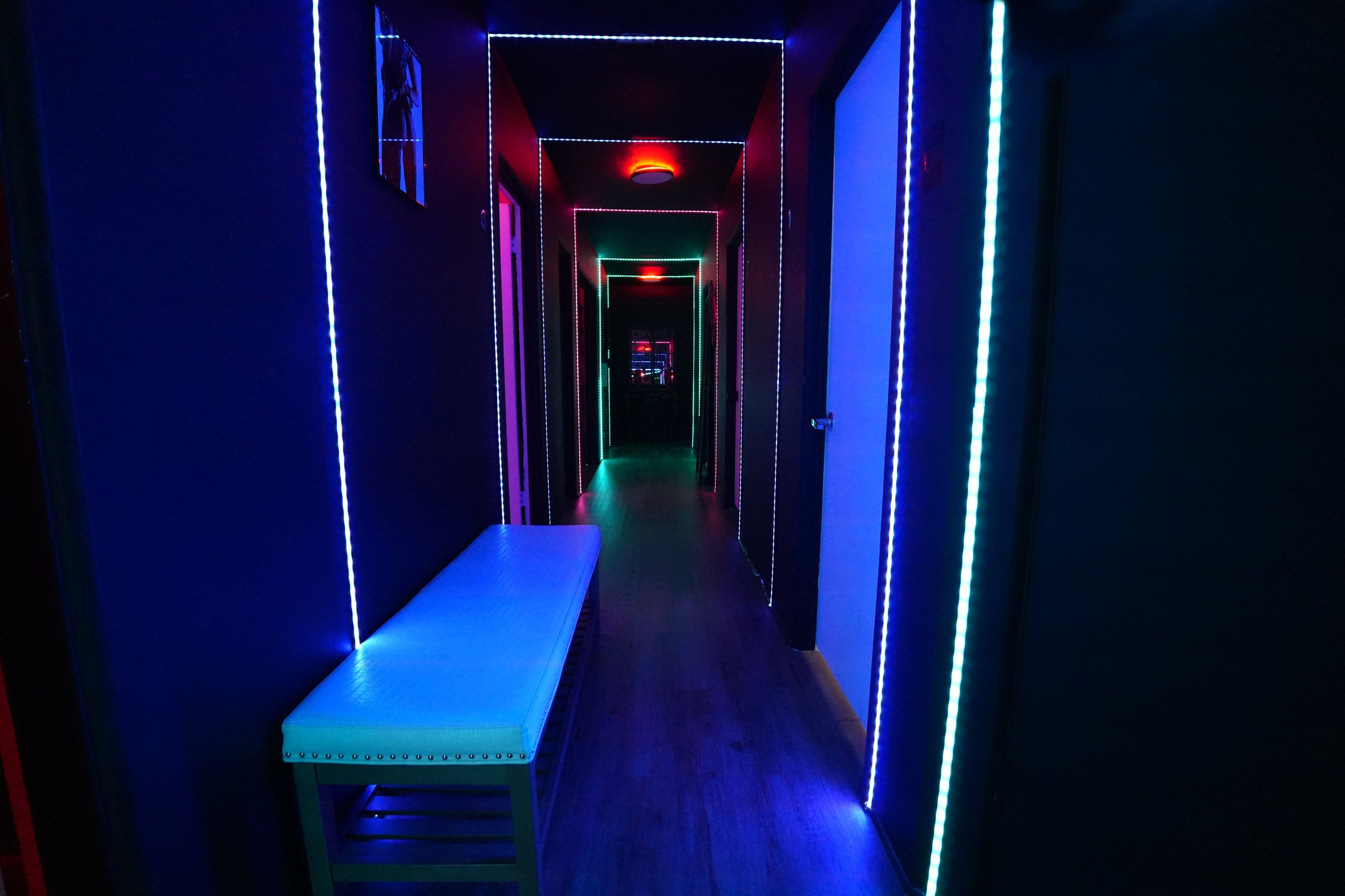 The image shows a narrow hallway illuminated with colorful LED lights along the edges and featuring a white bench on one side.
