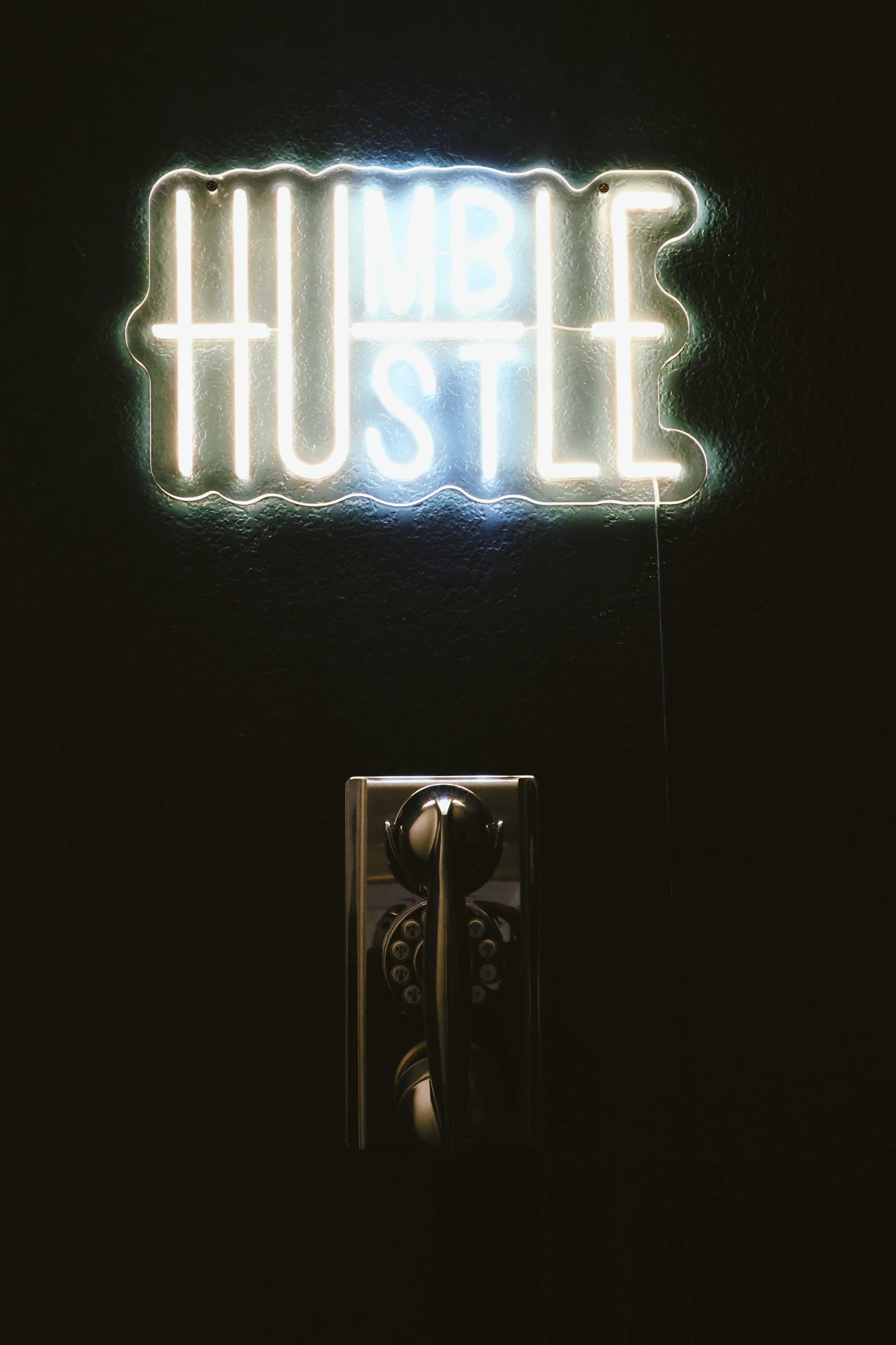 A neon sign reading "HUMBLE HUSTLE" is illuminated on a dark wall above an old-fashioned rotary telephone.