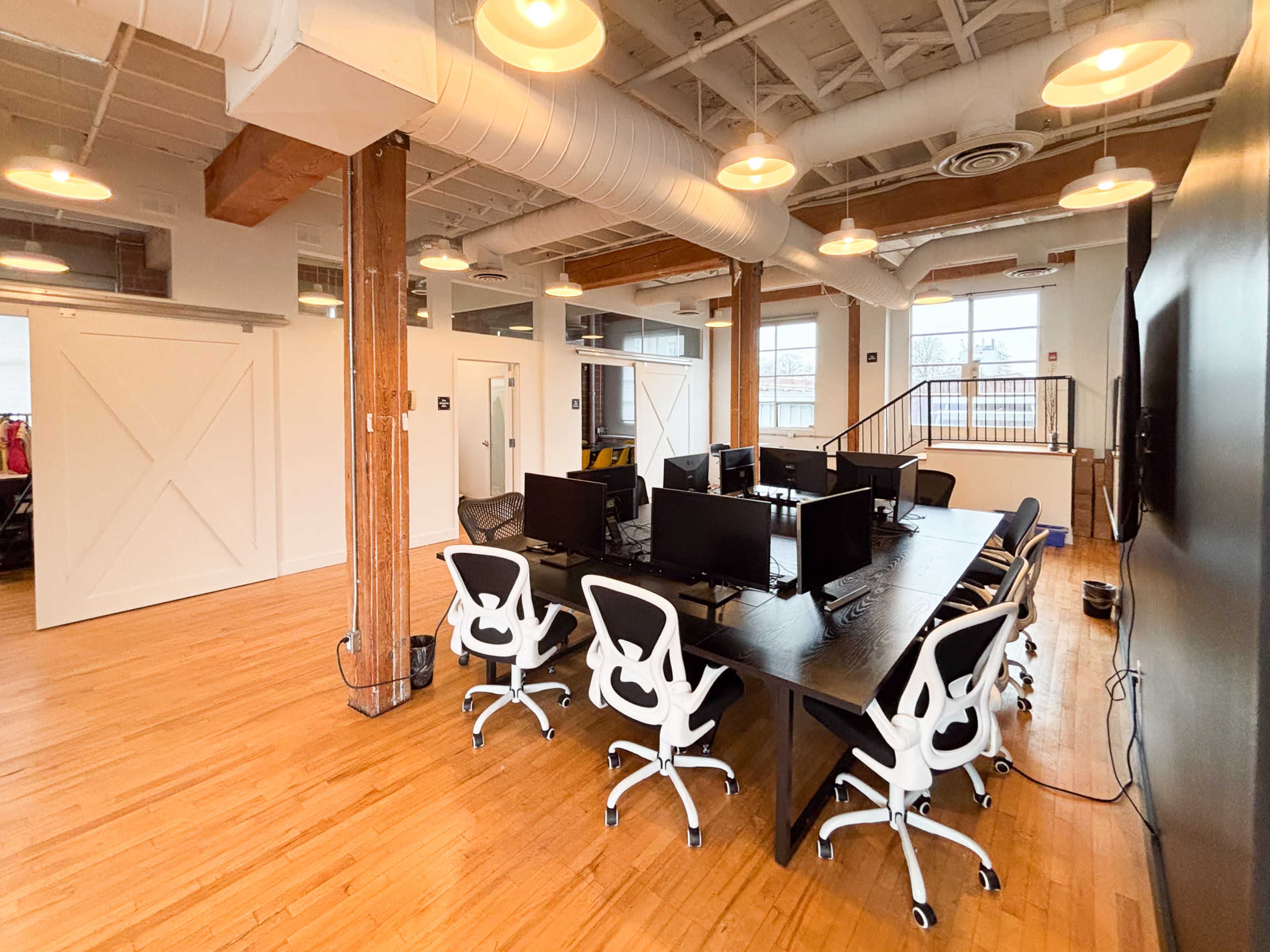 The image shows a modern office space with multiple black desks and white ergonomic chairs arranged in a well-lit, open area featuring wooden flooring and exposed beams.