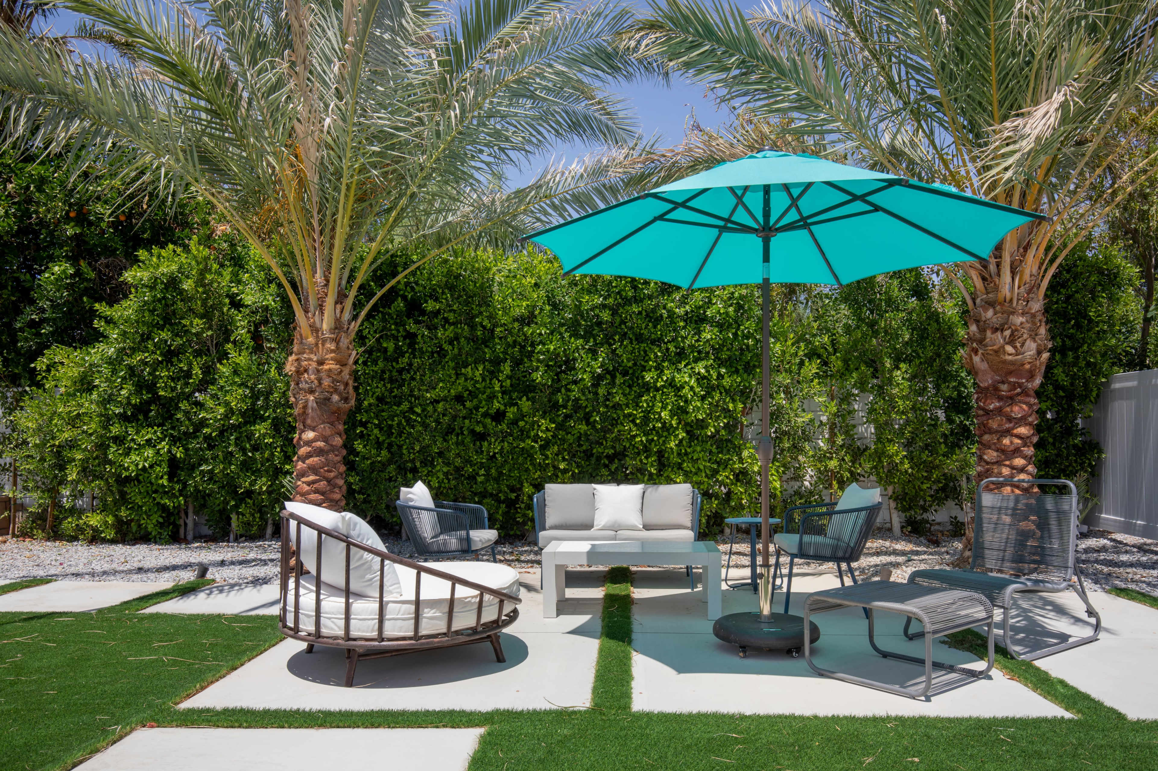 A seating area with white furniture and turquoise umbrellas is surrounded by palm trees and green grass.