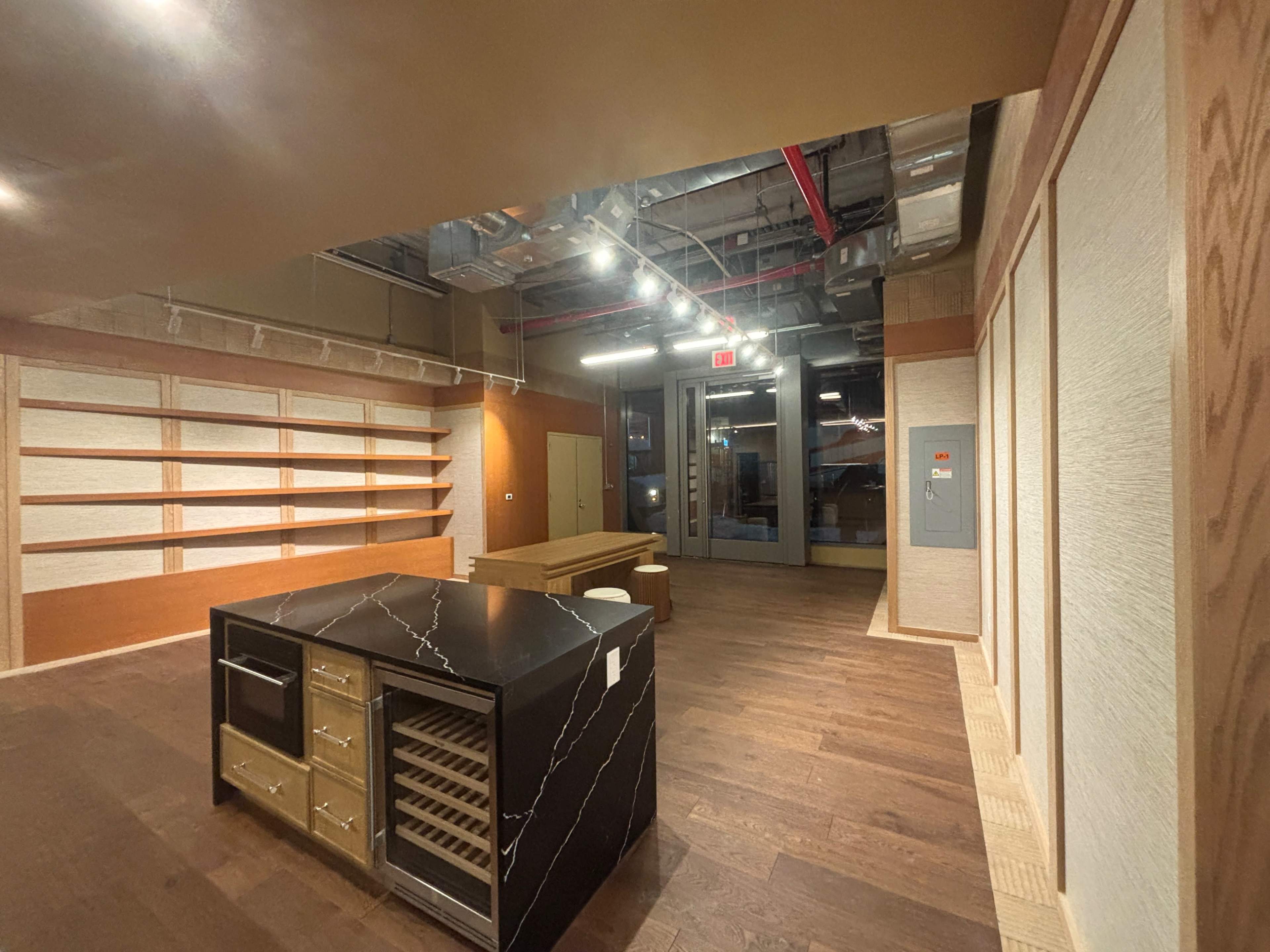 The image shows an empty retail space featuring wooden floors, a black marble counter with storage, and wall shelving units.