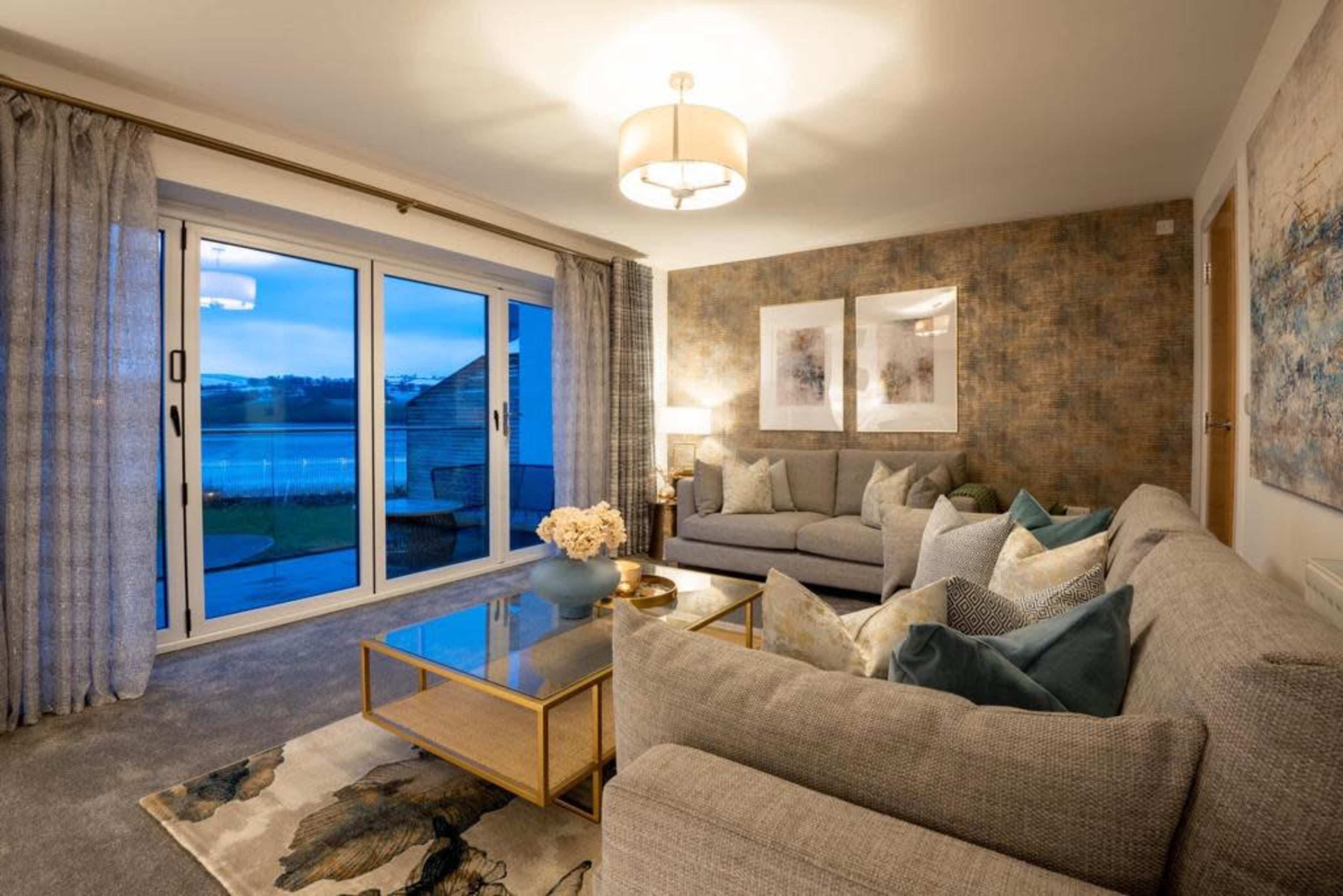 The image depicts a modern living room with gray sofas, a glass coffee table, and large windows that offer a view of the outdoors.