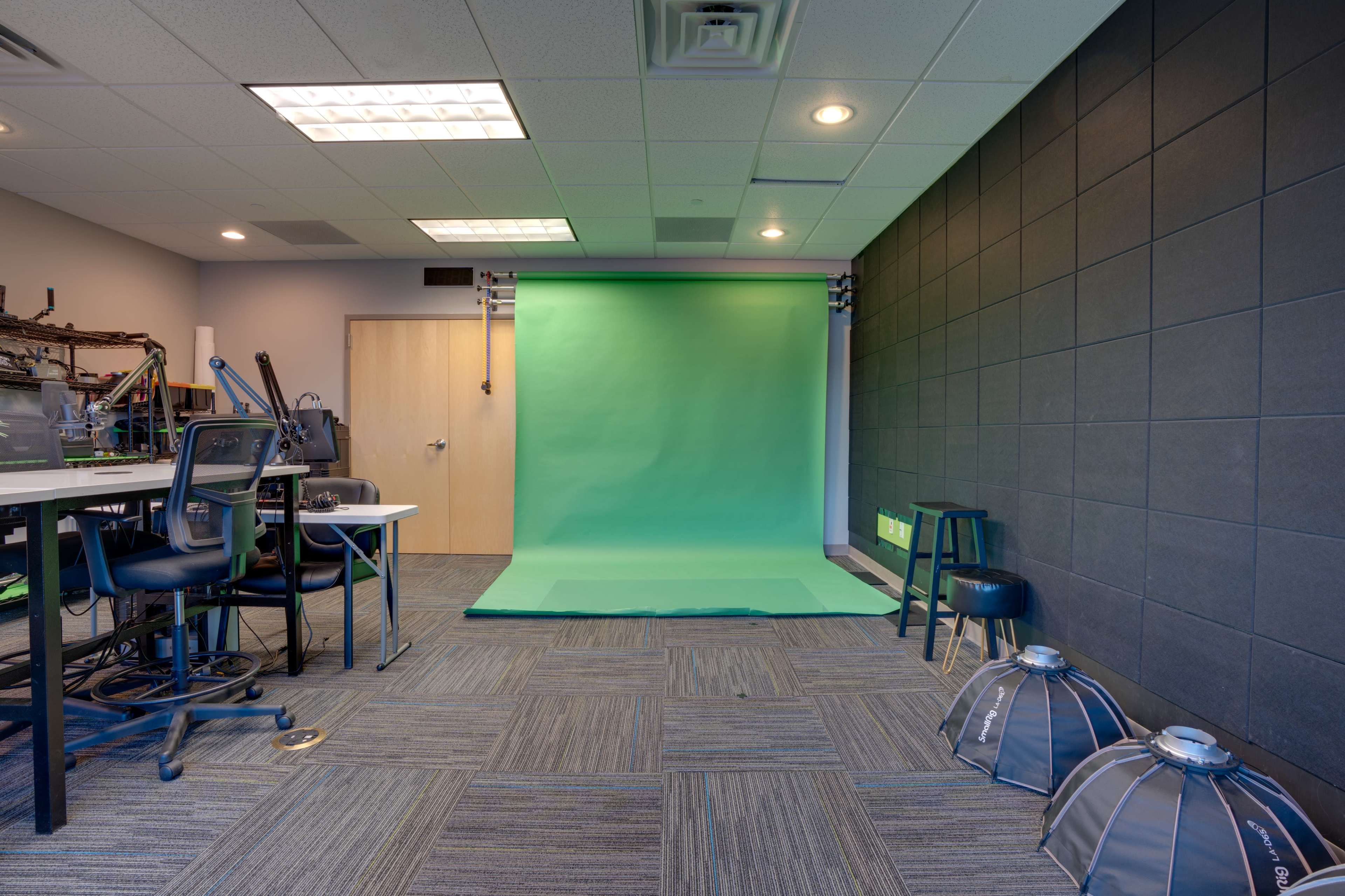 A room with a green backdrop and studio lights, accompanied by chairs and equipment on one side.