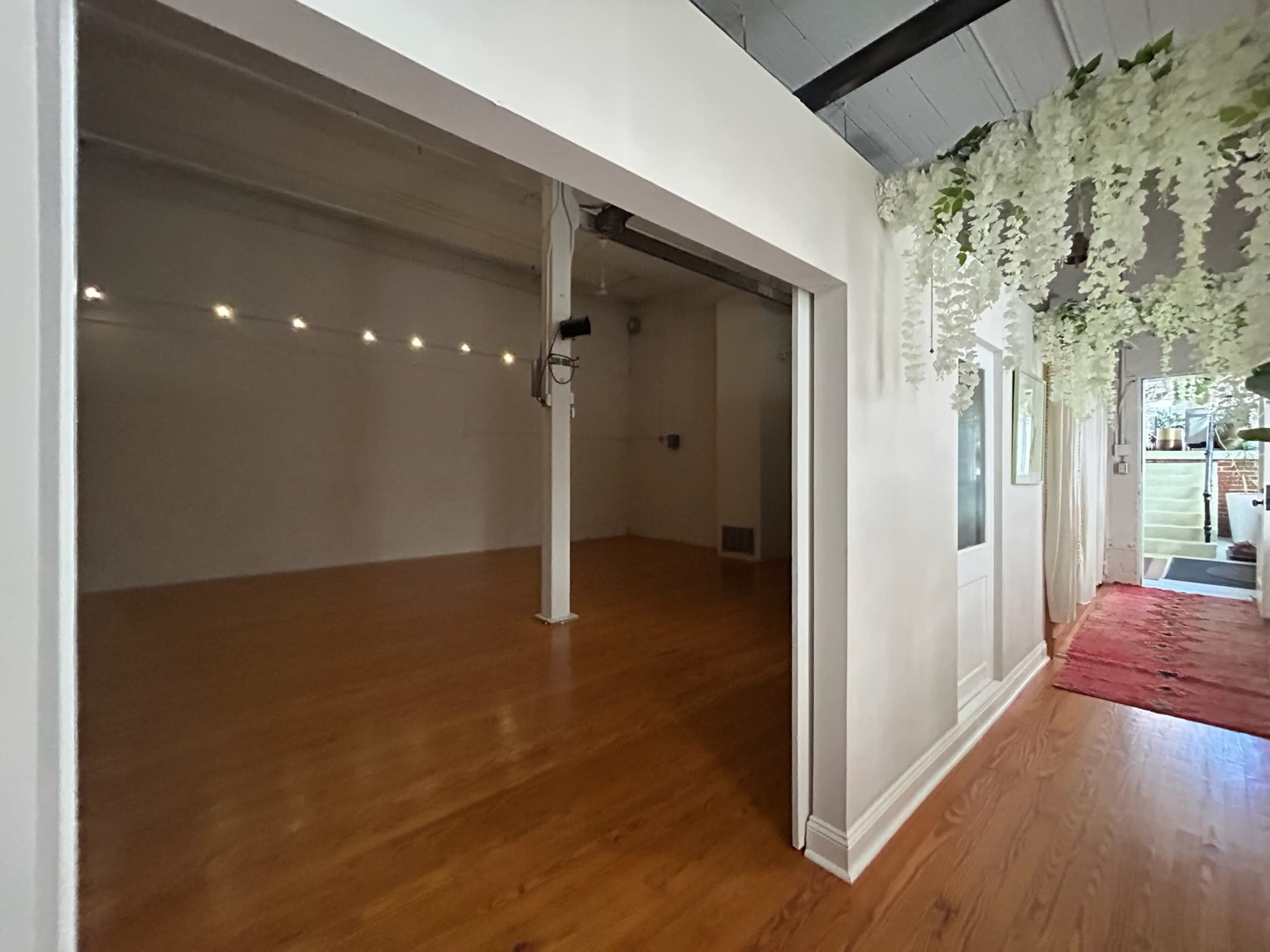 The image shows a spacious room with wooden flooring, featuring a large opening to another area decorated with hanging flowers.