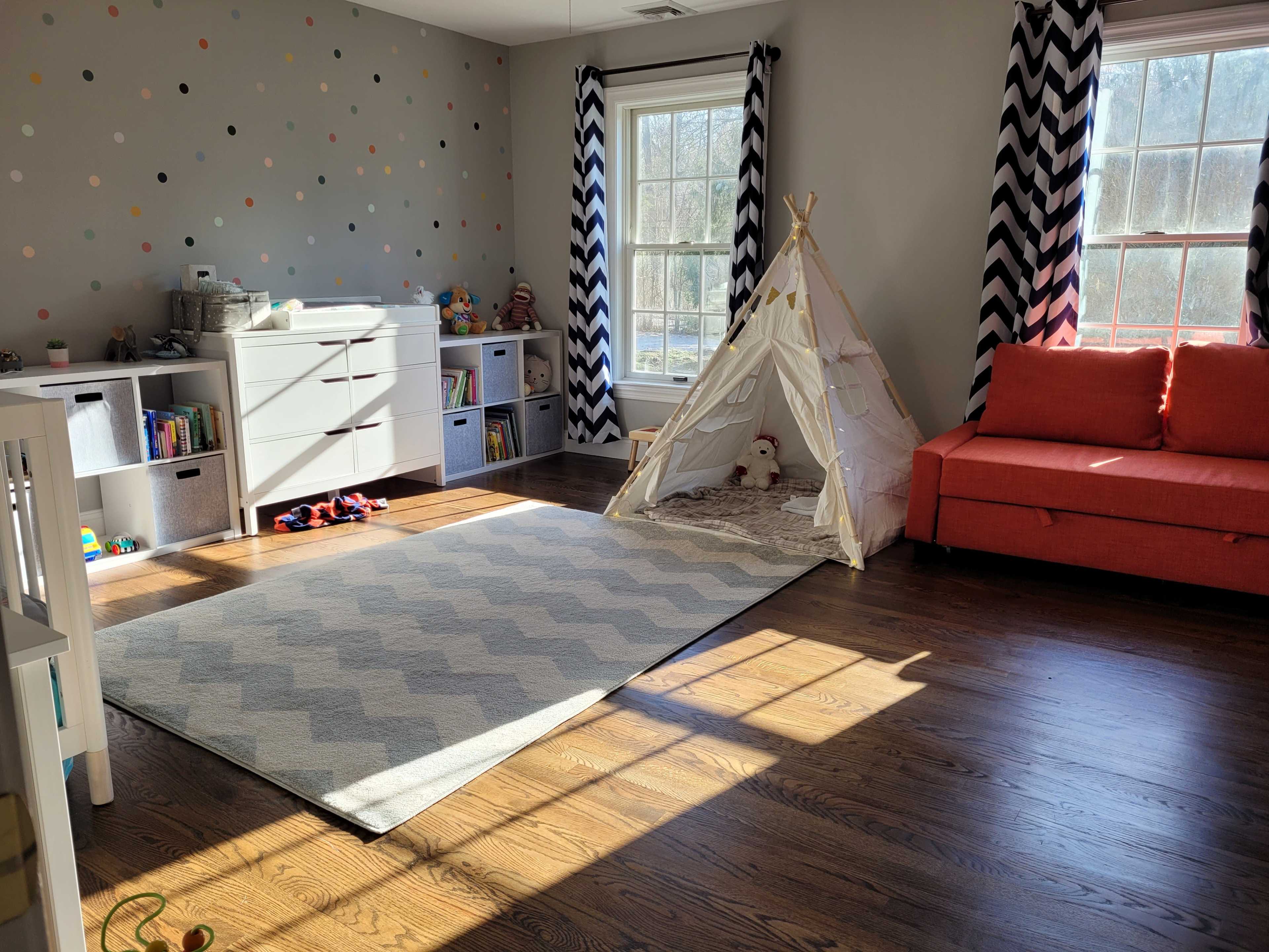 The room features a playful design with a sofa, a teepee tent, and organized storage, illuminated by natural light through large windows.