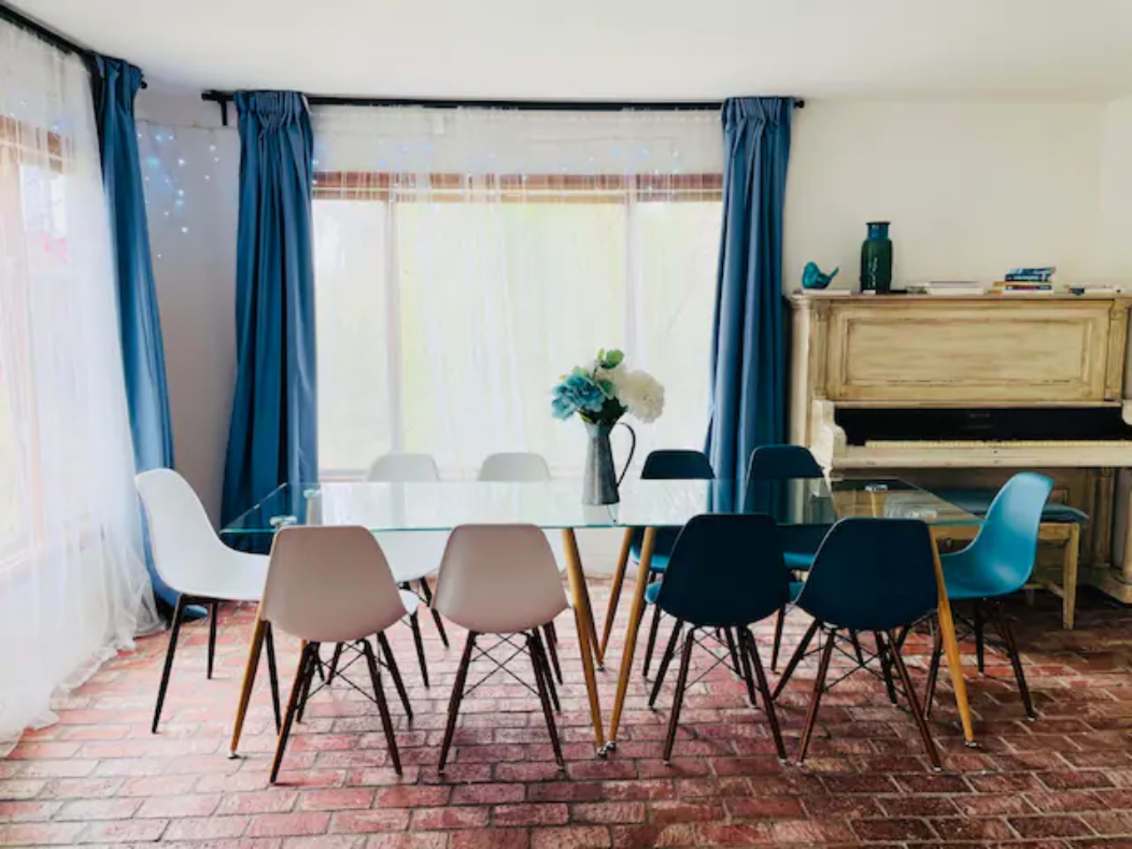 A bright dining room features a glass table surrounded by colorful chairs, with large windows draped in blue curtains and a piano in the corner.