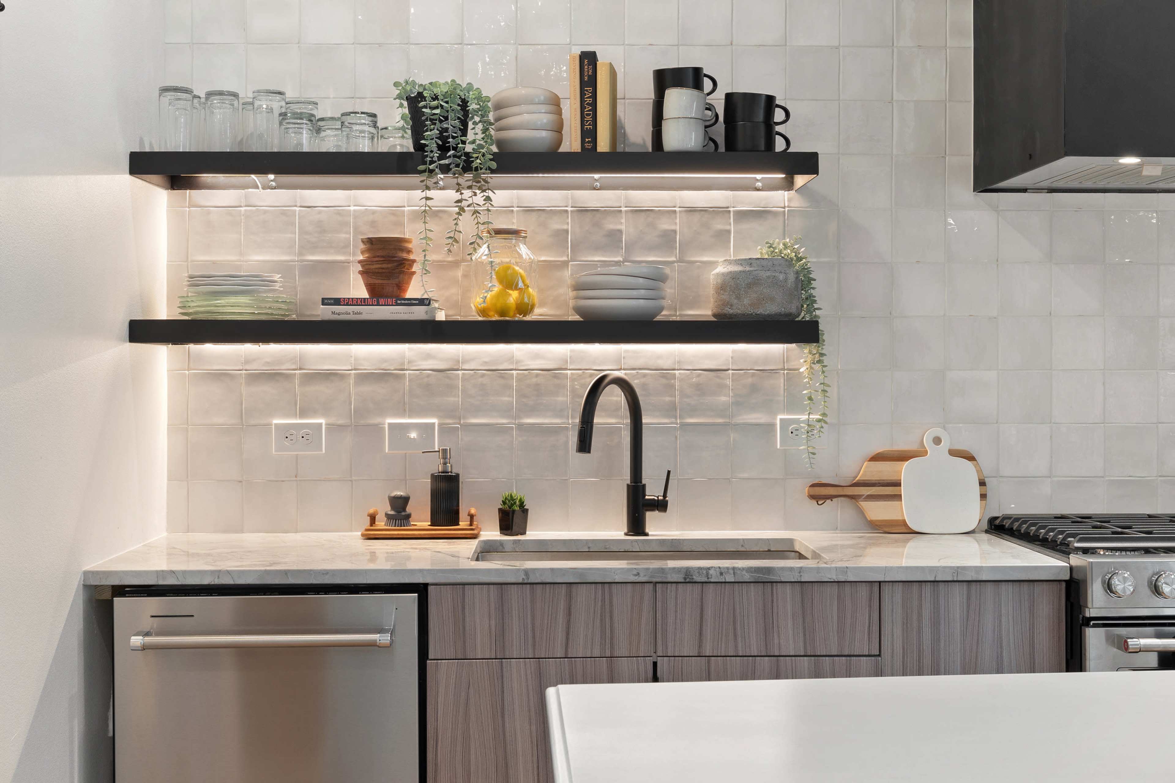 A modern kitchen with two floating shelves above a marble countertop, featuring a sink, a dishwasher, and various kitchen items.