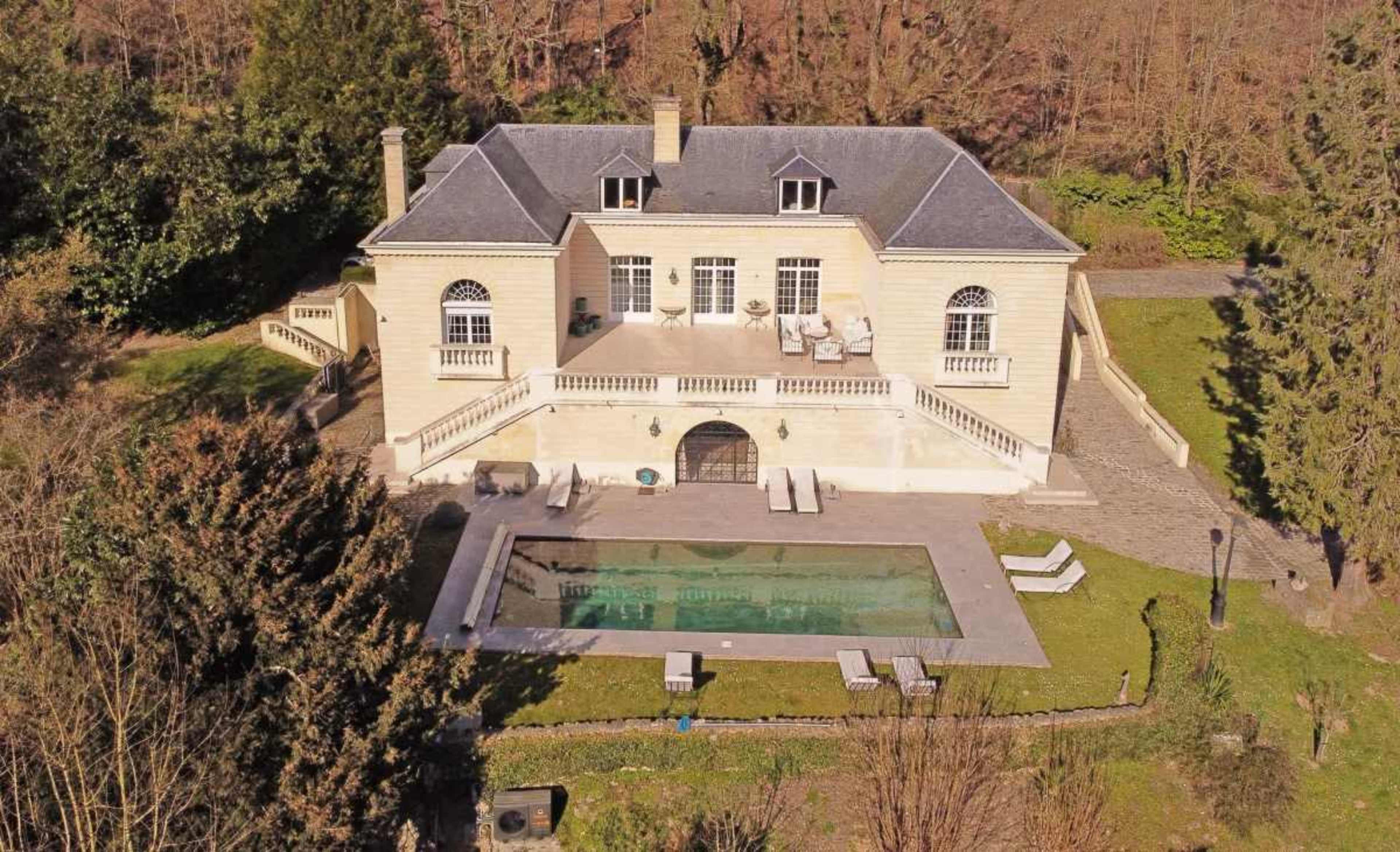 The image shows a large, two-story mansion with a pool in the backyard, surrounded by trees and greenery.