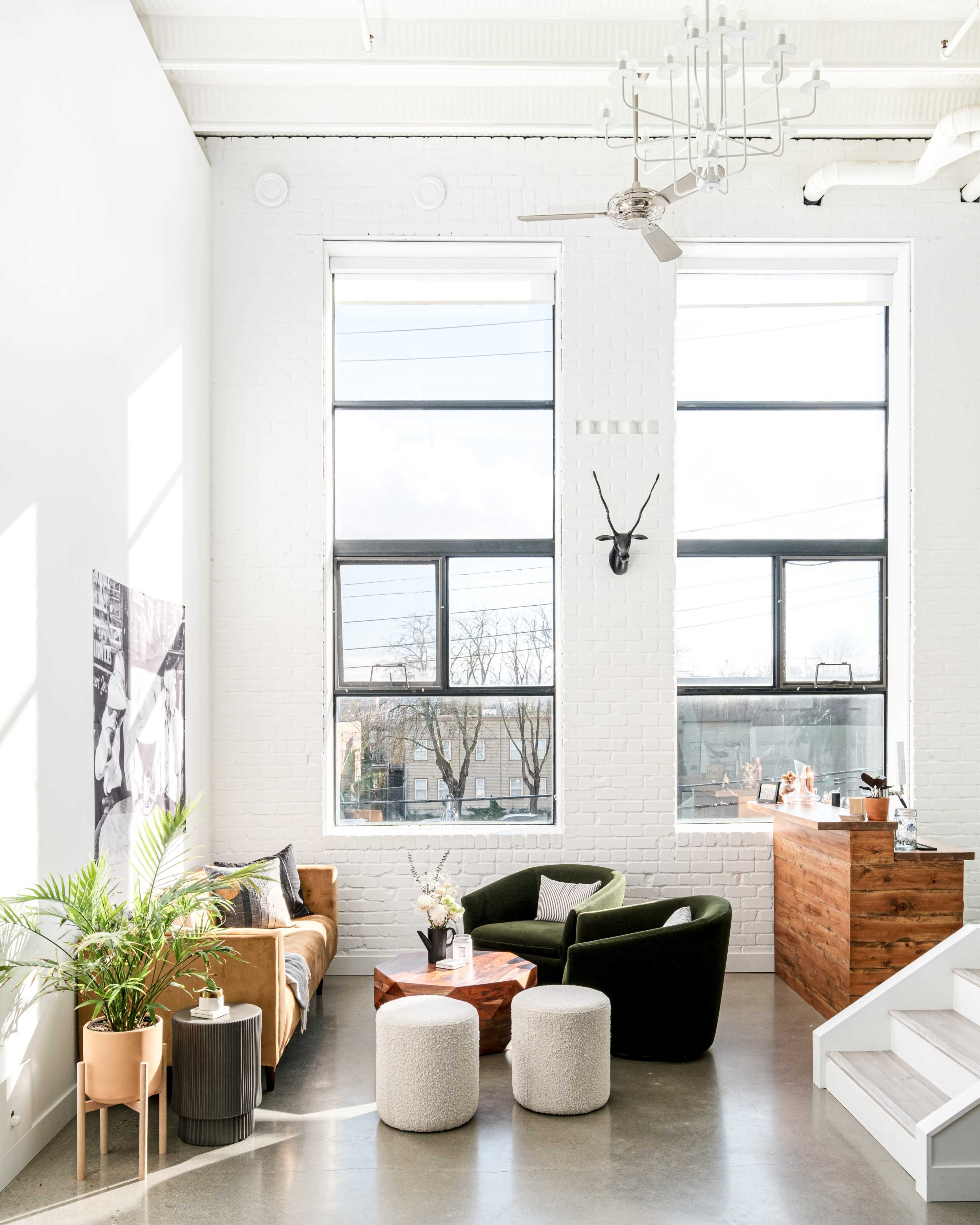 A bright, modern living space with large windows, a seating area featuring a brown sofa and green chairs, and a wooden coffee table surrounded by light-colored stools.
