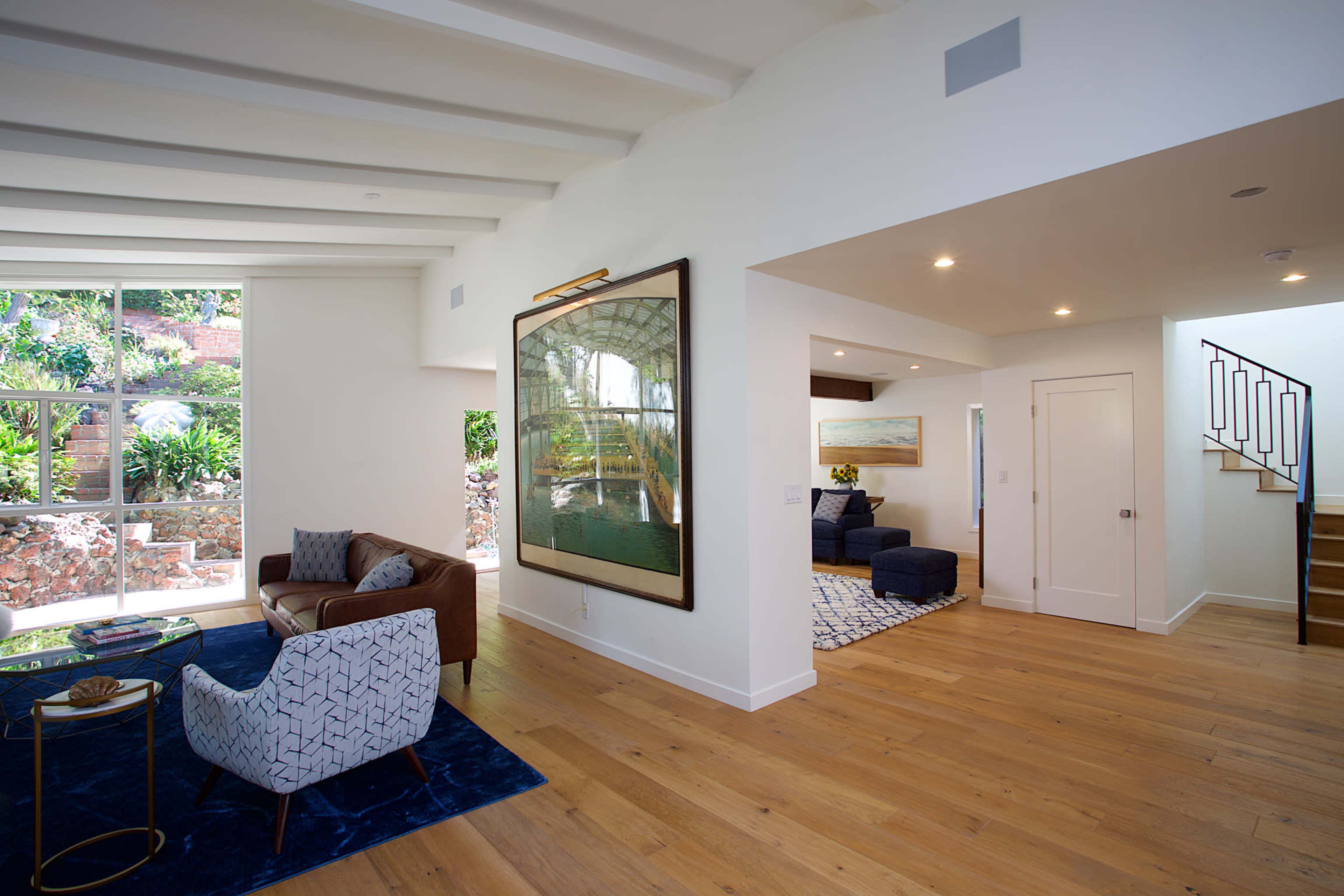 A modern living space with a leather sofa, a patterned chair, and a staircase, featuring large windows that allow natural light and provide a view of greenery outside.