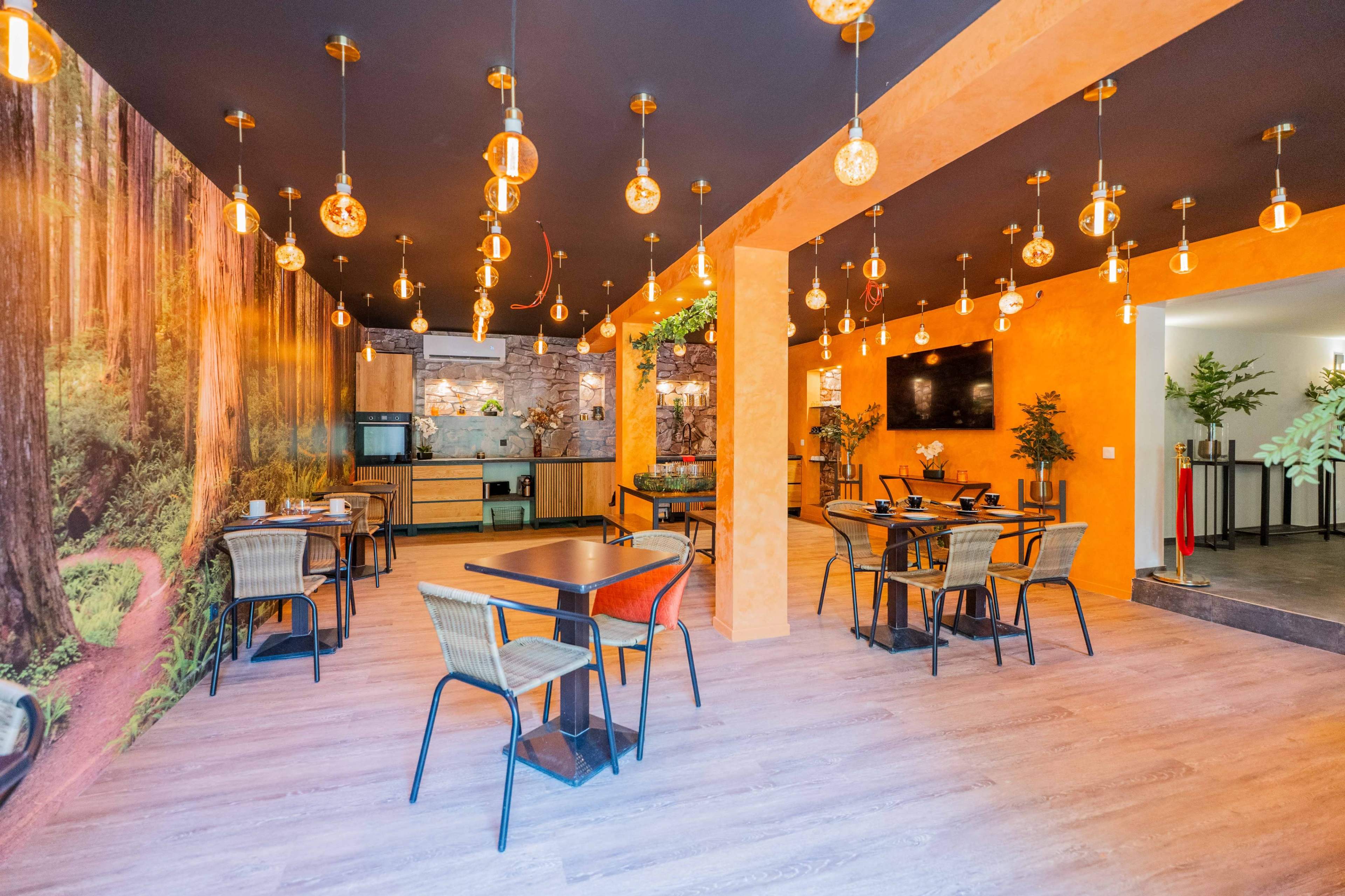 The image shows a modern cafe interior with wooden flooring, orange walls, and a mural of a forest, featuring several tables and hanging light fixtures.