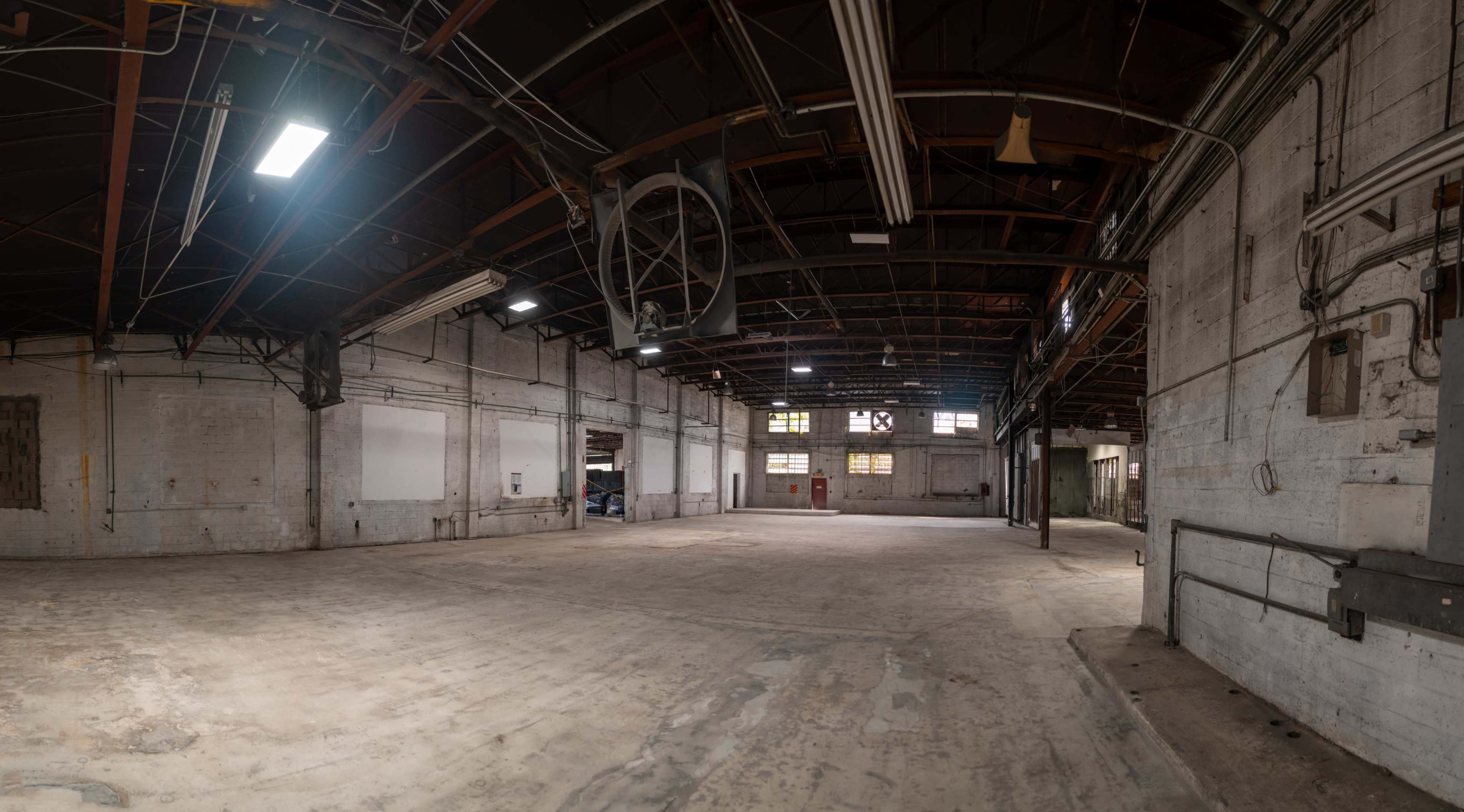 The image shows a spacious, empty warehouse with high ceilings, exposed beams, and concrete floors, illuminated by overhead lights.