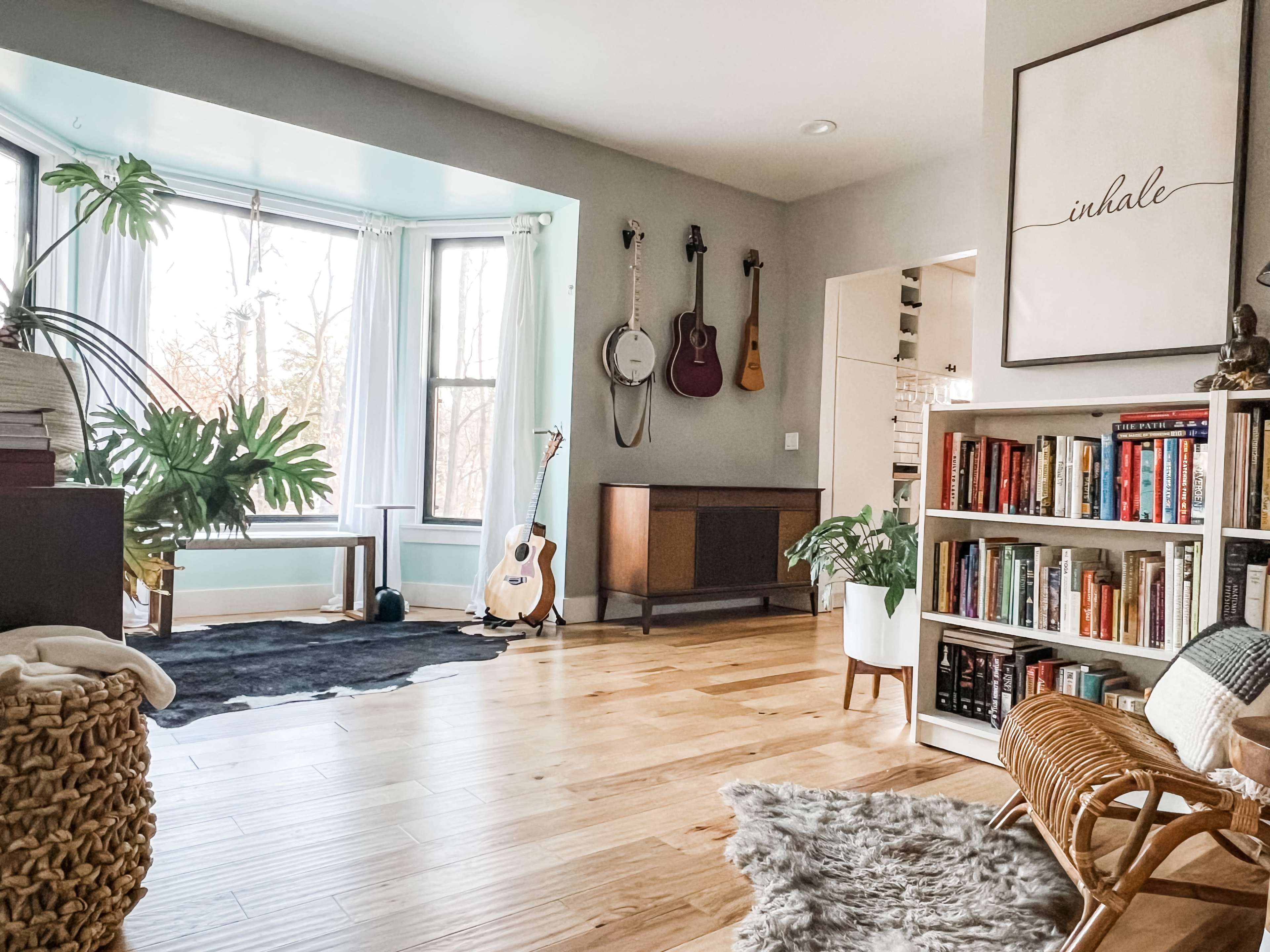 The image shows a spacious living room featuring large windows, a bookshelf filled with books, an acoustic guitar on a stand, and a cozy chair placed next to a decorative plant.