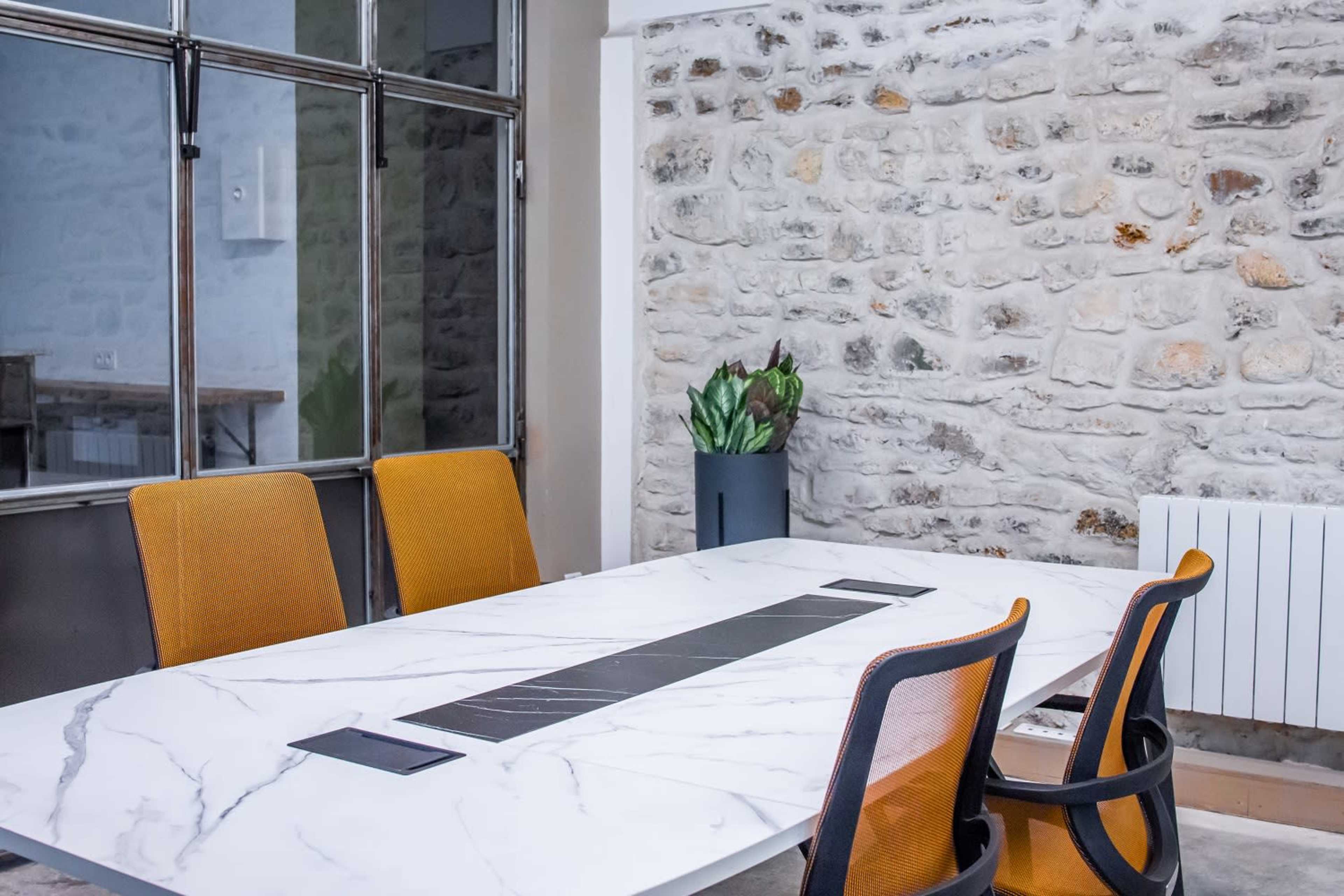 A conference room features a marble table with orange chairs, set against a stone wall and large windows.