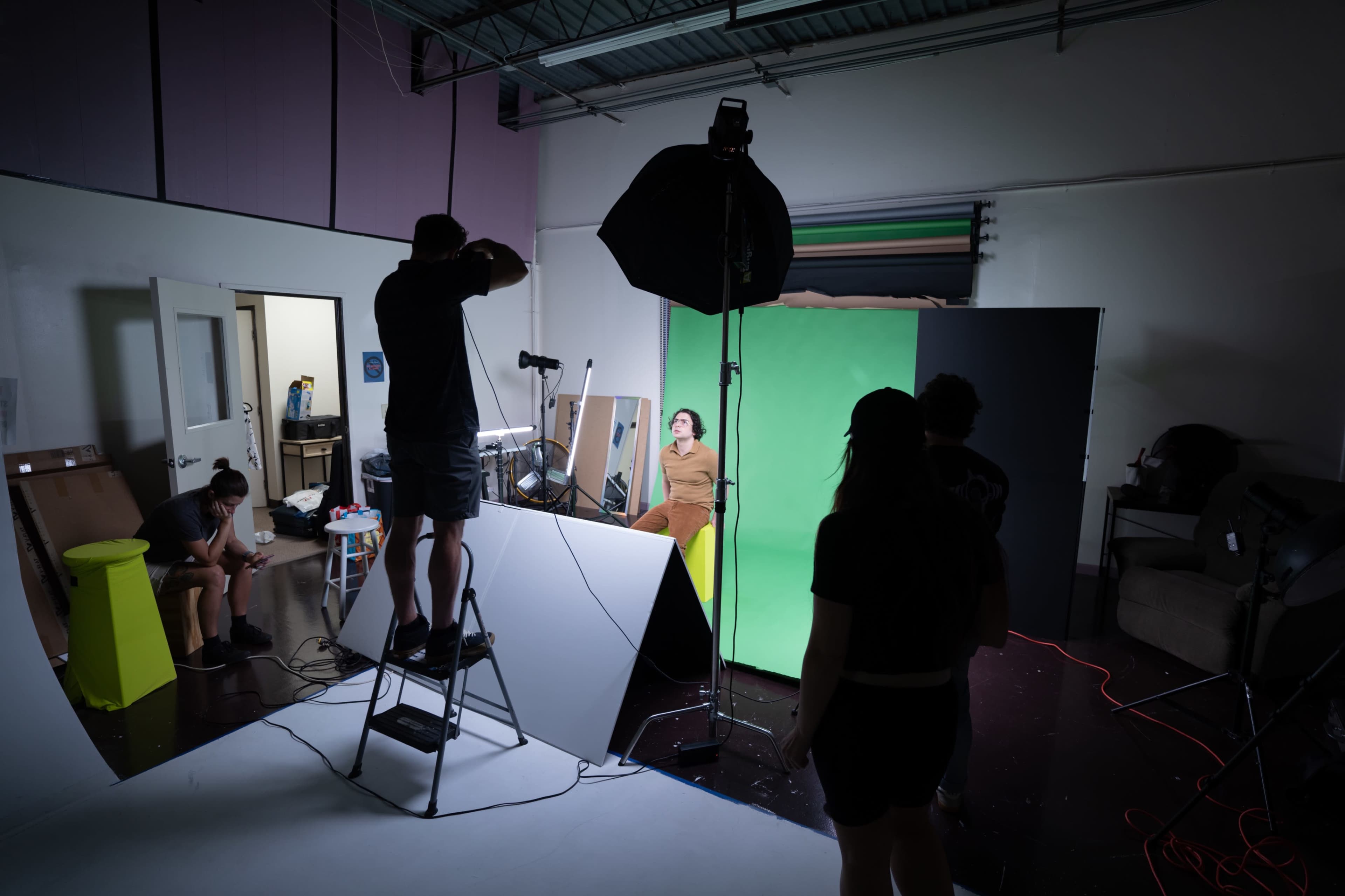 A photography studio is set up with a model posing on a set, while photographers and assistants adjust lighting and equipment around them.
