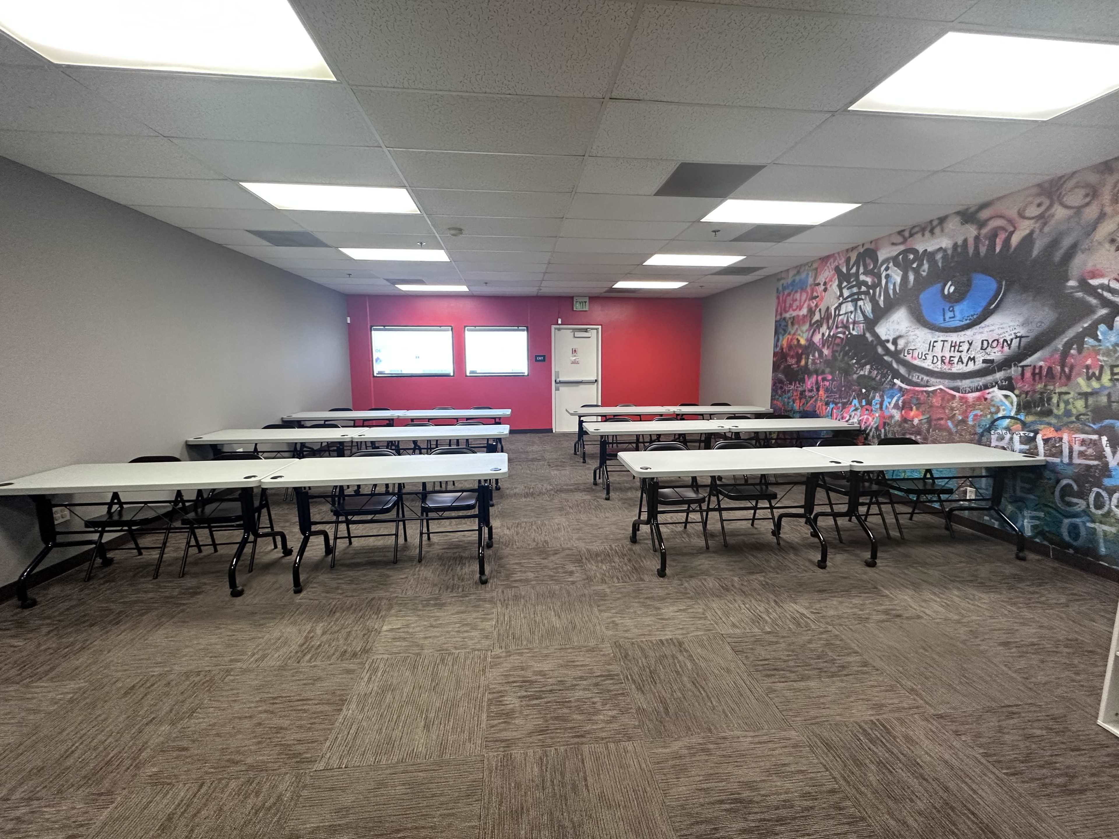 A classroom or meeting room with several white folding tables arranged in rows, and a colorful graffiti mural on one wall.