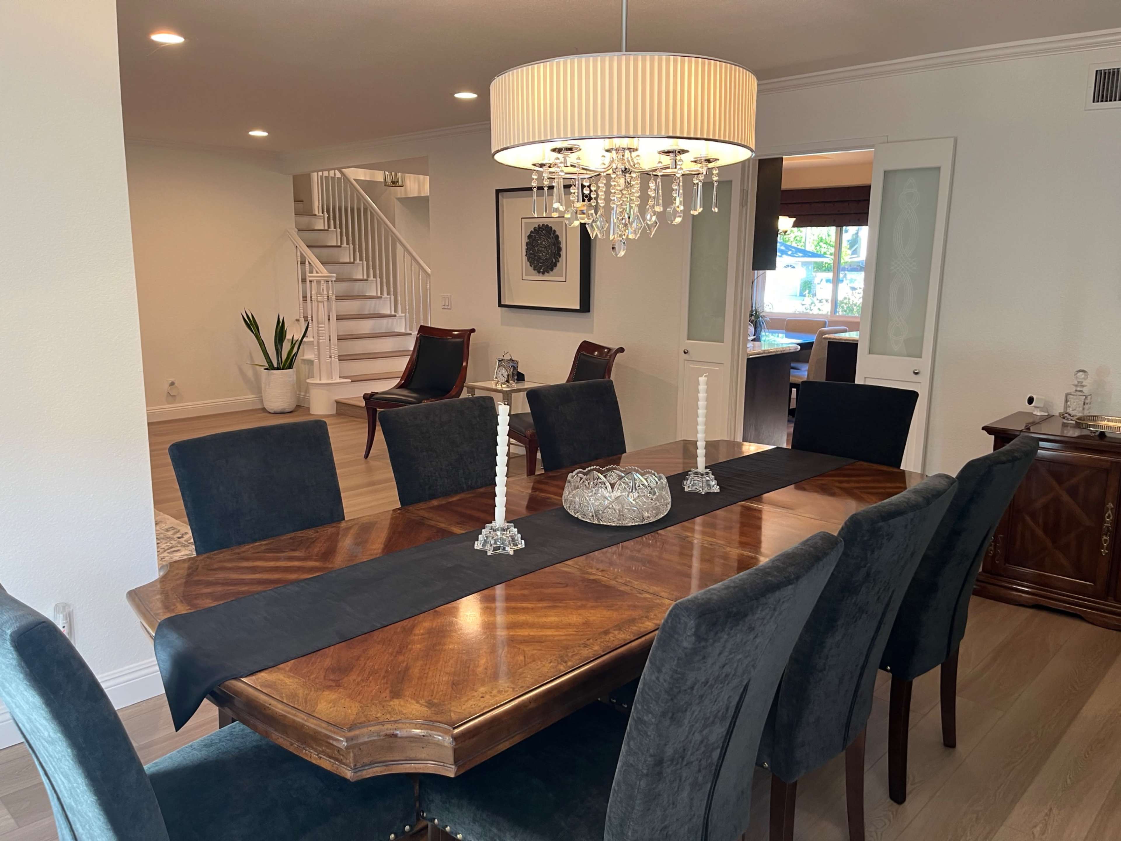 The image shows a modern dining room featuring a large wooden table surrounded by dark upholstered chairs, with a chandelier overhead and a staircase visible in the background.