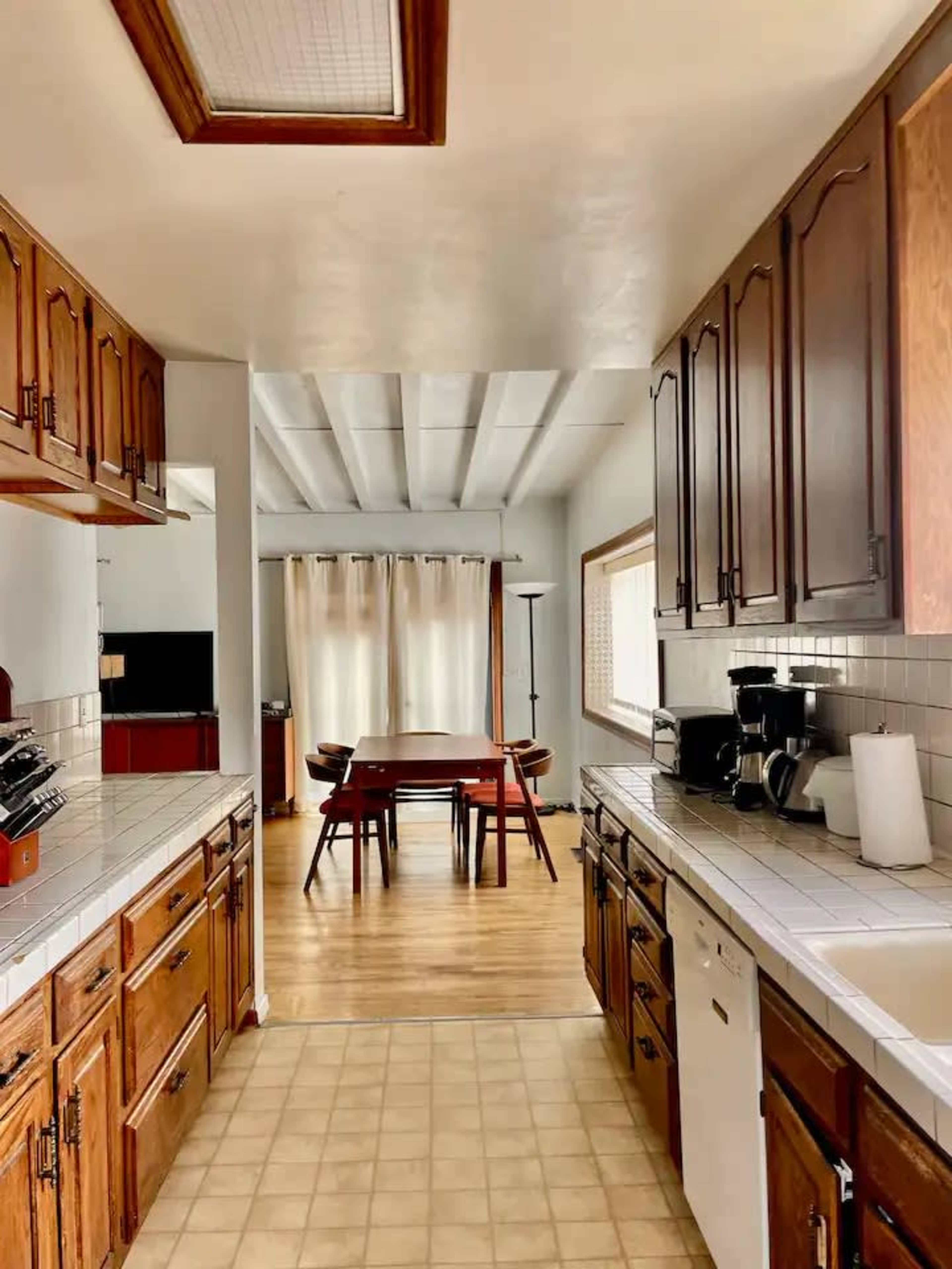 A kitchen with wooden cabinets and white countertops leads to a dining area, which features a table surrounded by chairs and large windows with sheer curtains.