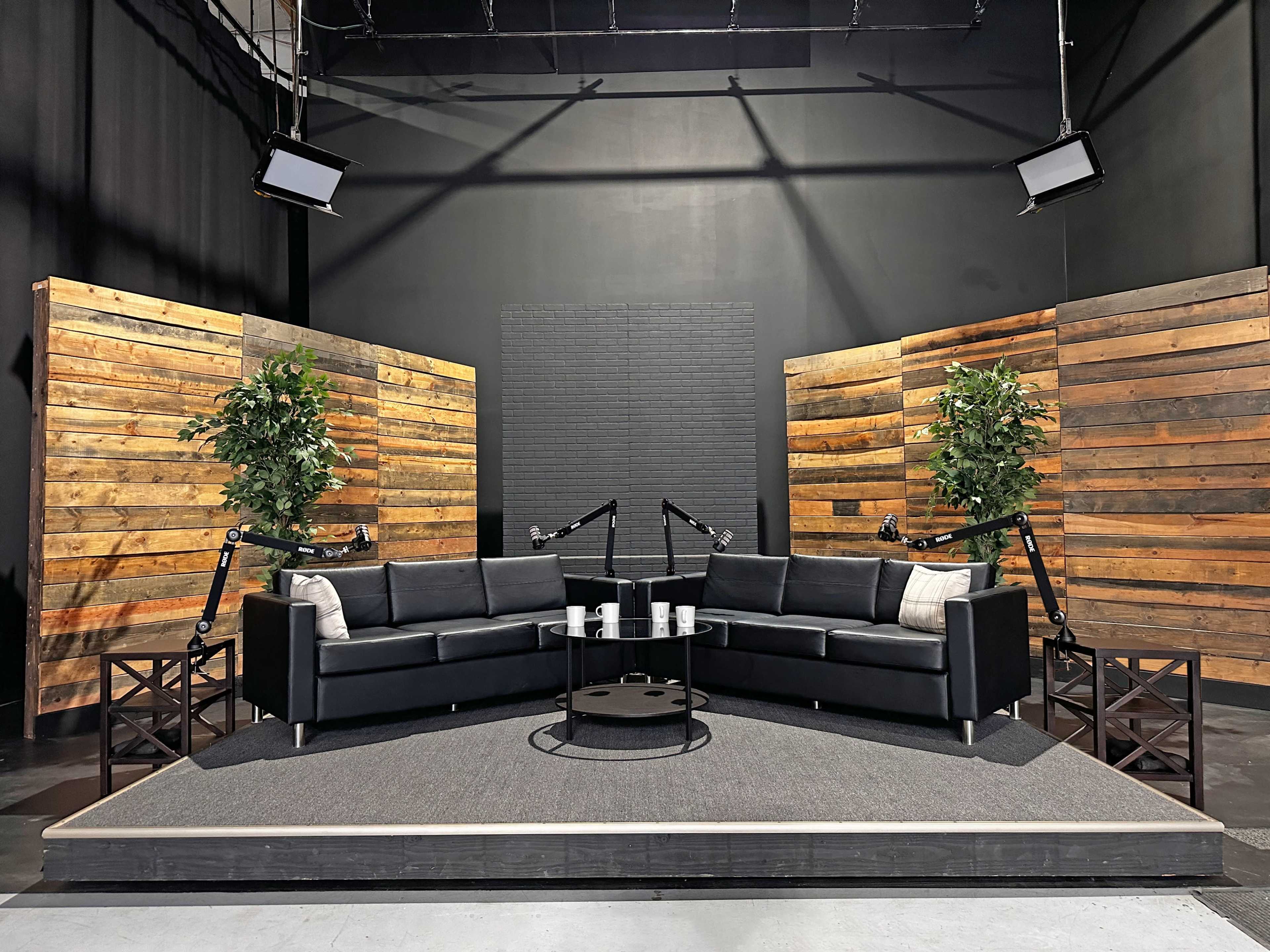 The image shows a television studio set featuring a black sofa arrangement on a raised platform, surrounded by wooden panel backdrops and two potted plants.