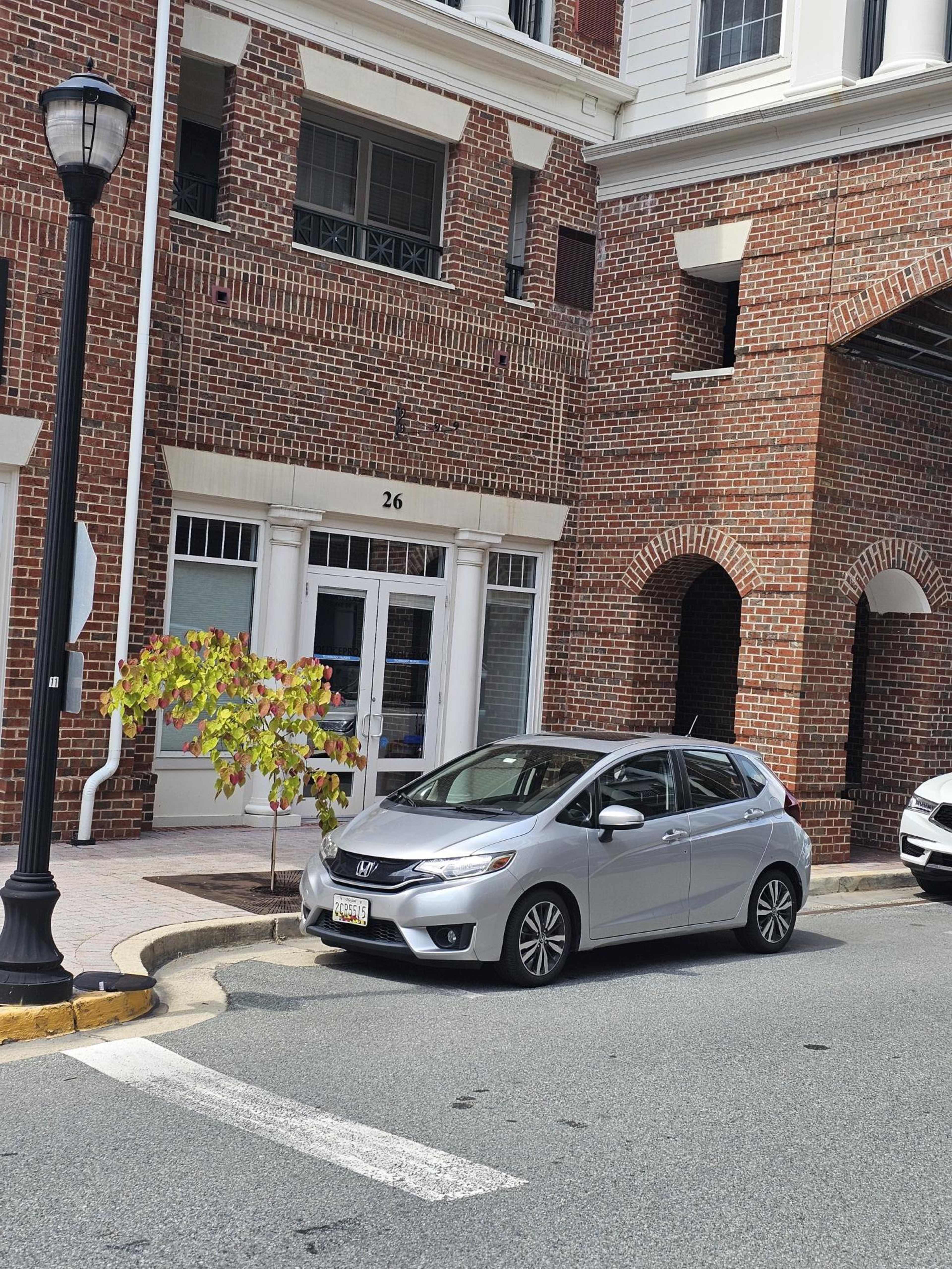 A silver Honda Fit is parked near a brick building with a number 26 on it.