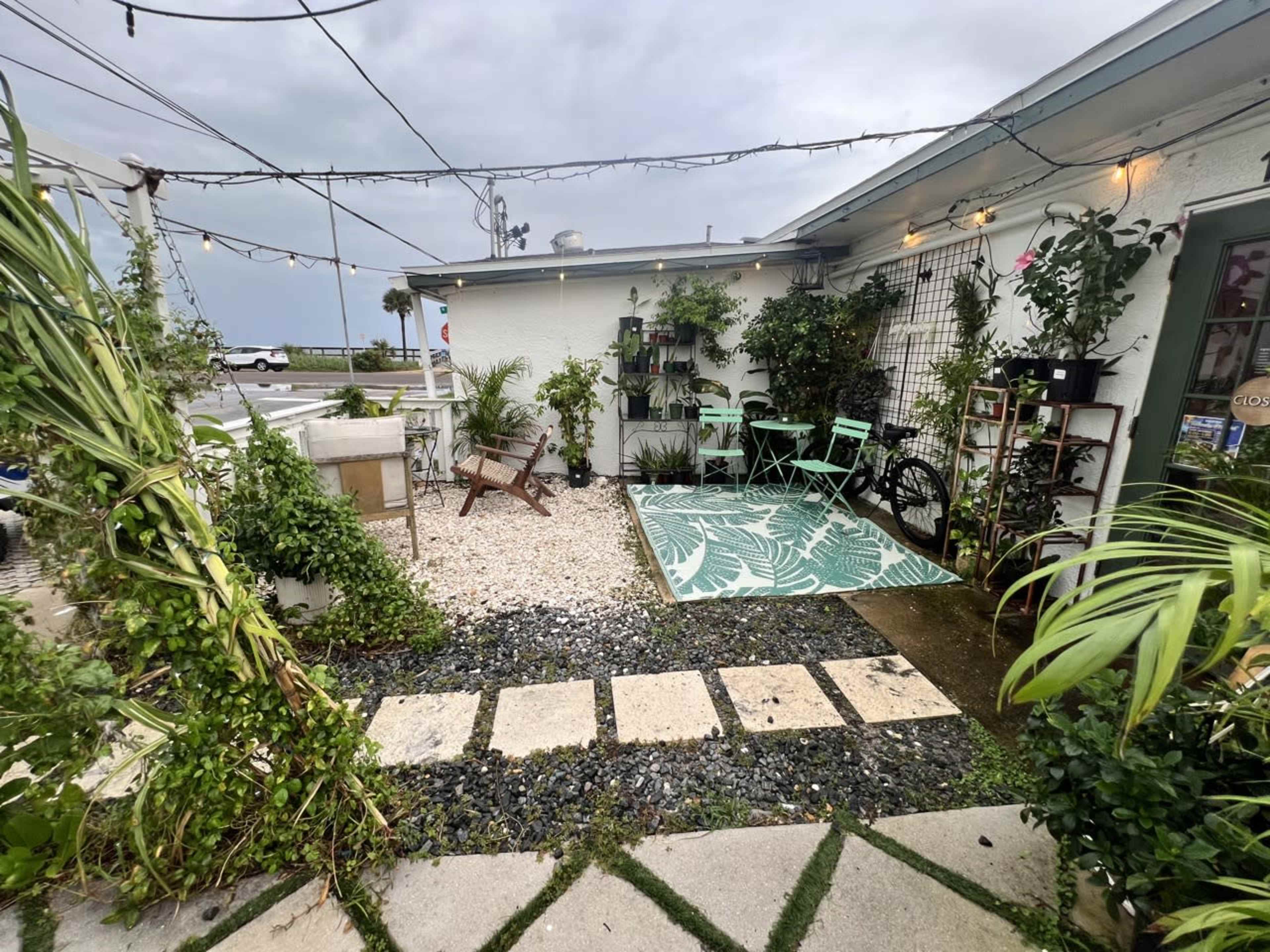 The image shows a small outdoor seating area with plants, stone pathways, and two green chairs next to a building.