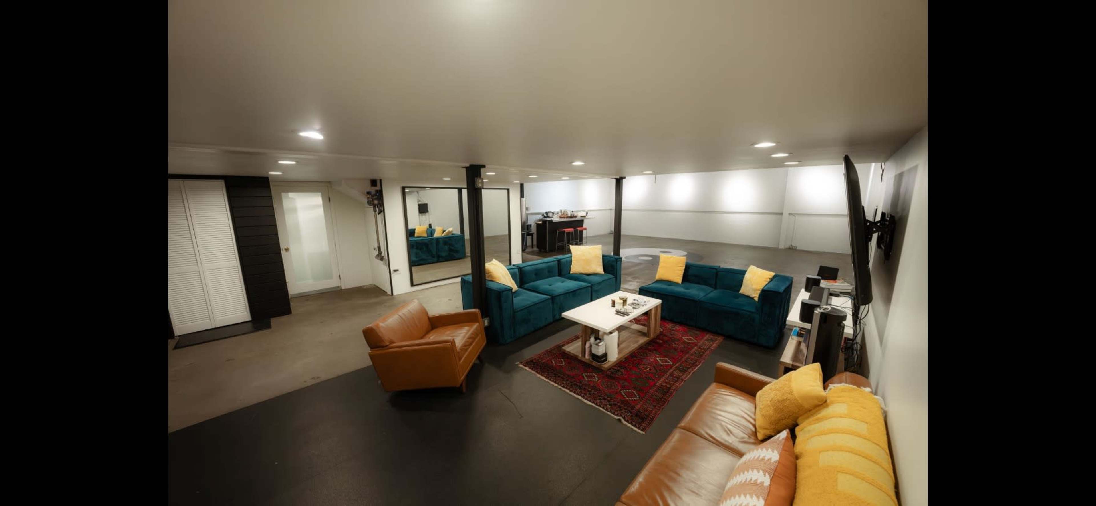 A spacious indoor area with a sectional couch, a coffee table, and a mix of beige and blue chairs arranged around a patterned rug, along with a wall-mounted TV.